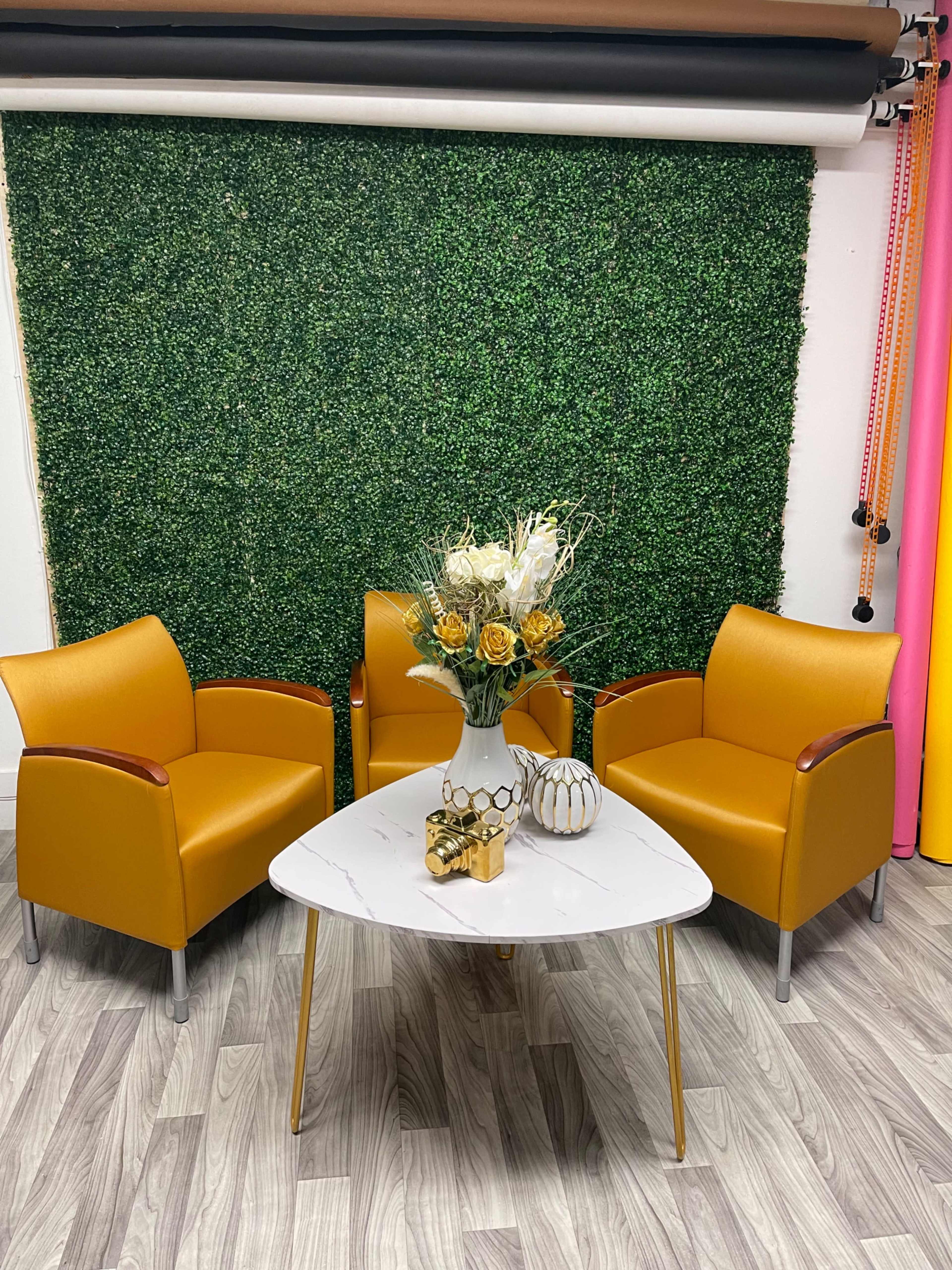 The scene features two mustard-colored chairs positioned around a white marble table with a floral arrangement and decorative items, set against a green hedge backdrop.