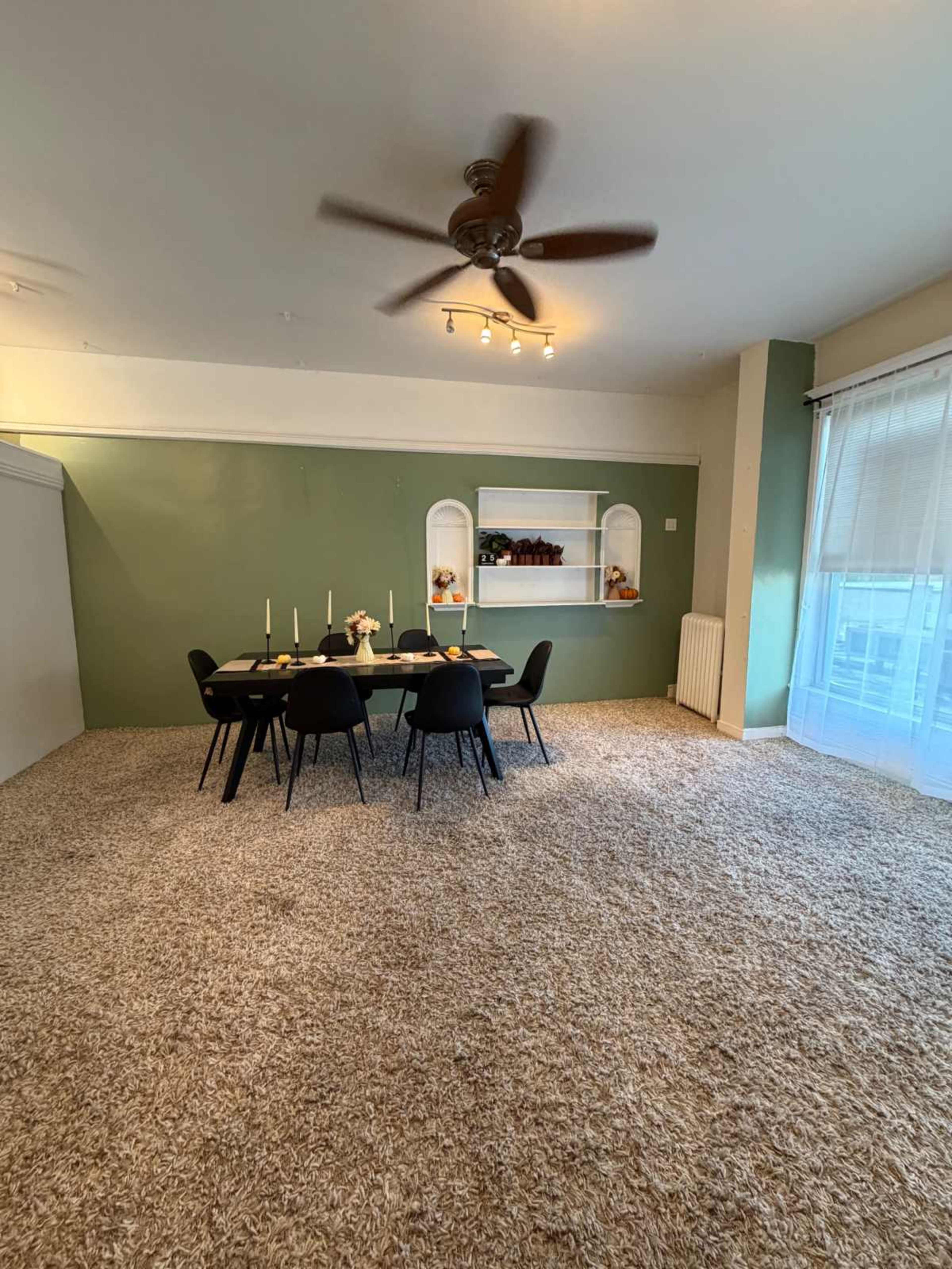 A spacious dining area features a round table with chairs, a wall-mounted shelf, and a ceiling fan, all on a carpeted floor.