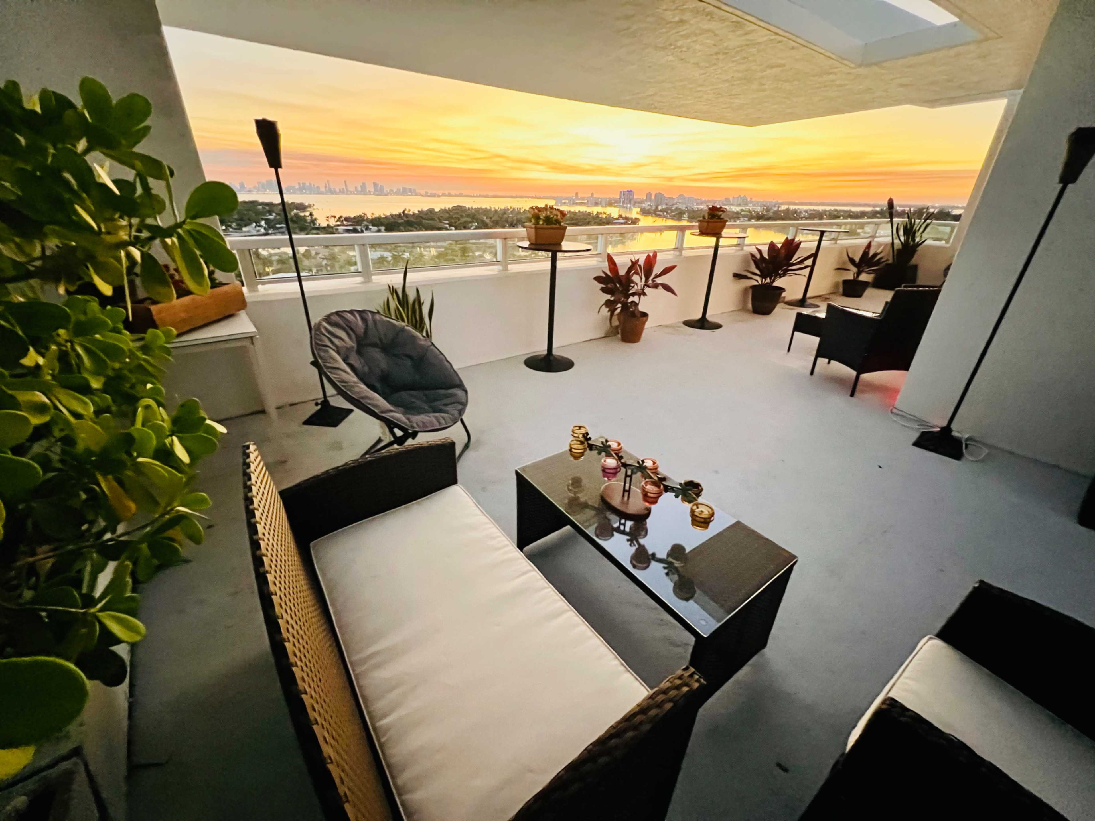 The image shows a balcony with seating furniture overlooking a scenic sunset view.