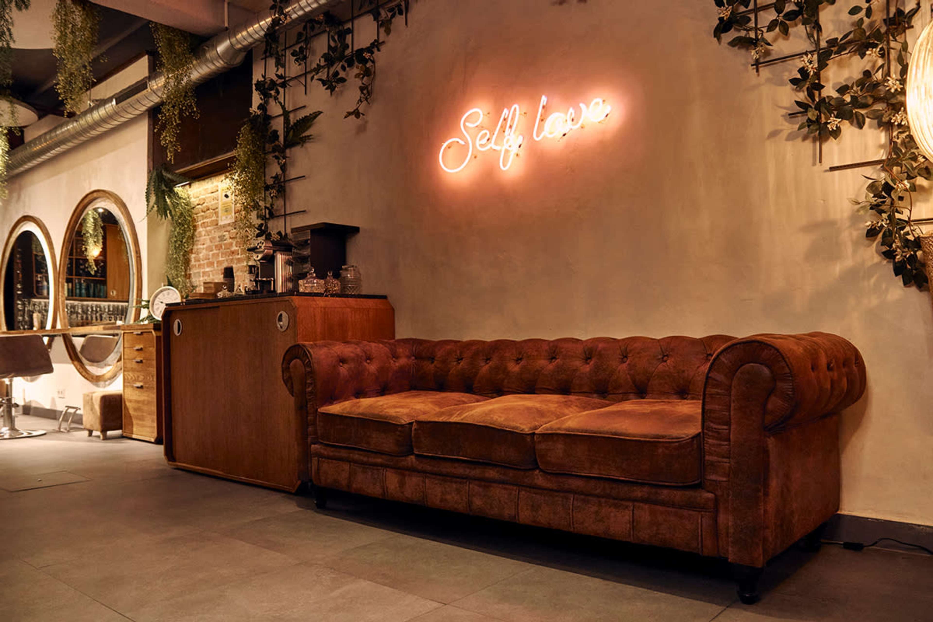 The scene features a brown leather couch against a decorated wall with a neon sign that reads "Self love."