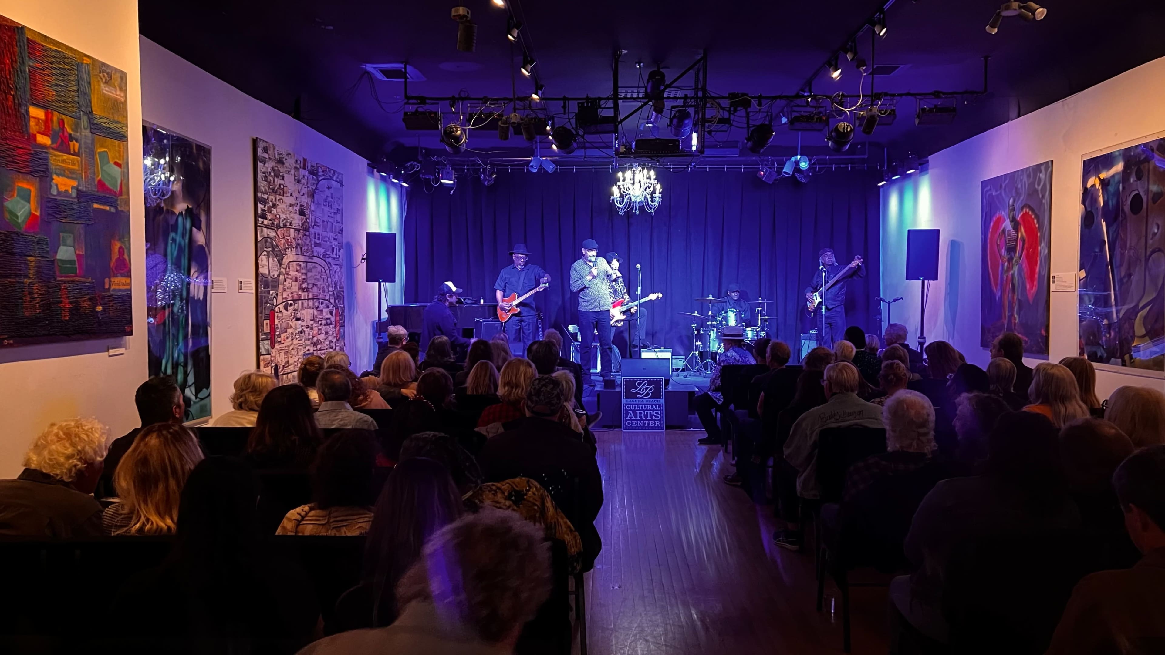 A live band performs on stage in a dimly lit gallery filled with seated audience members.