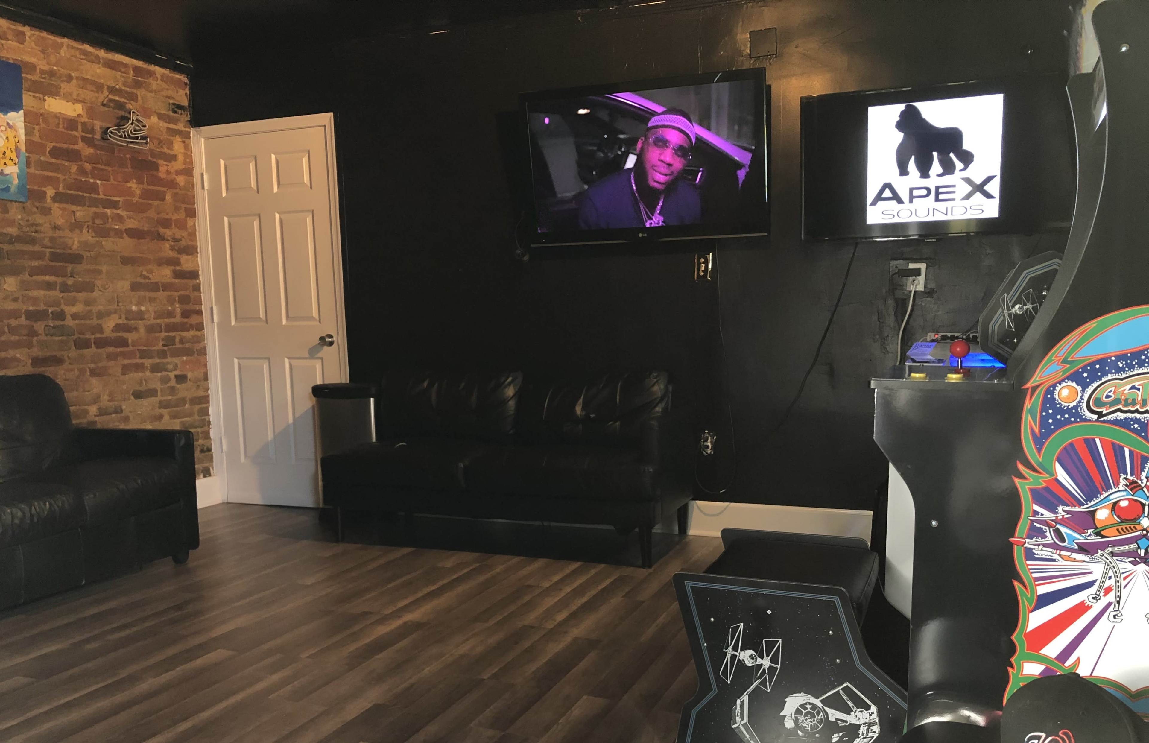 A room with a black wall, a leather couch, two televisions displaying a person and a logo, and an arcade game in the corner.