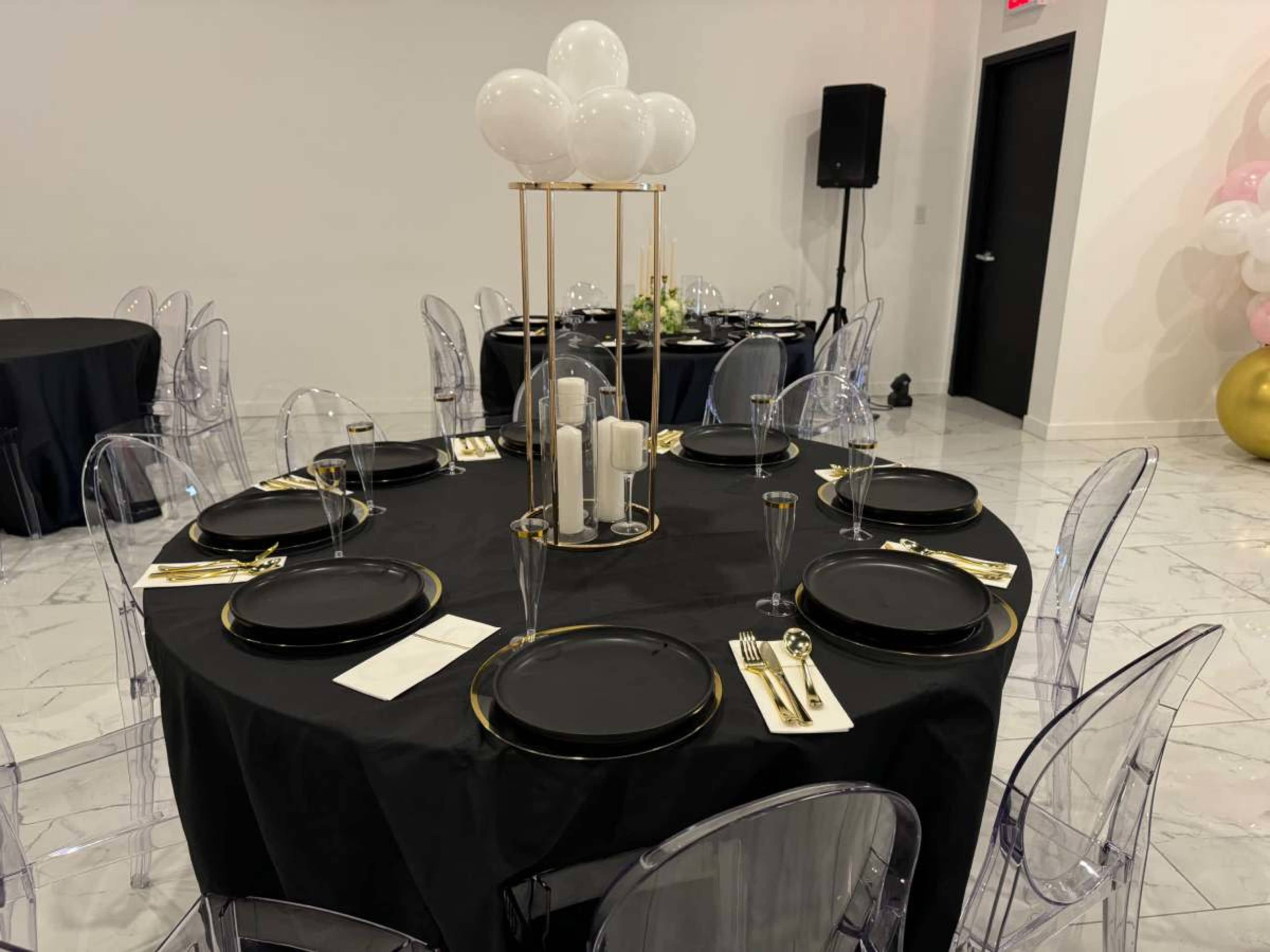 A round table is set with black tablecloth, gold-accented plates, clear glassware, and a centerpiece of candles and white balloons in a modern event space.