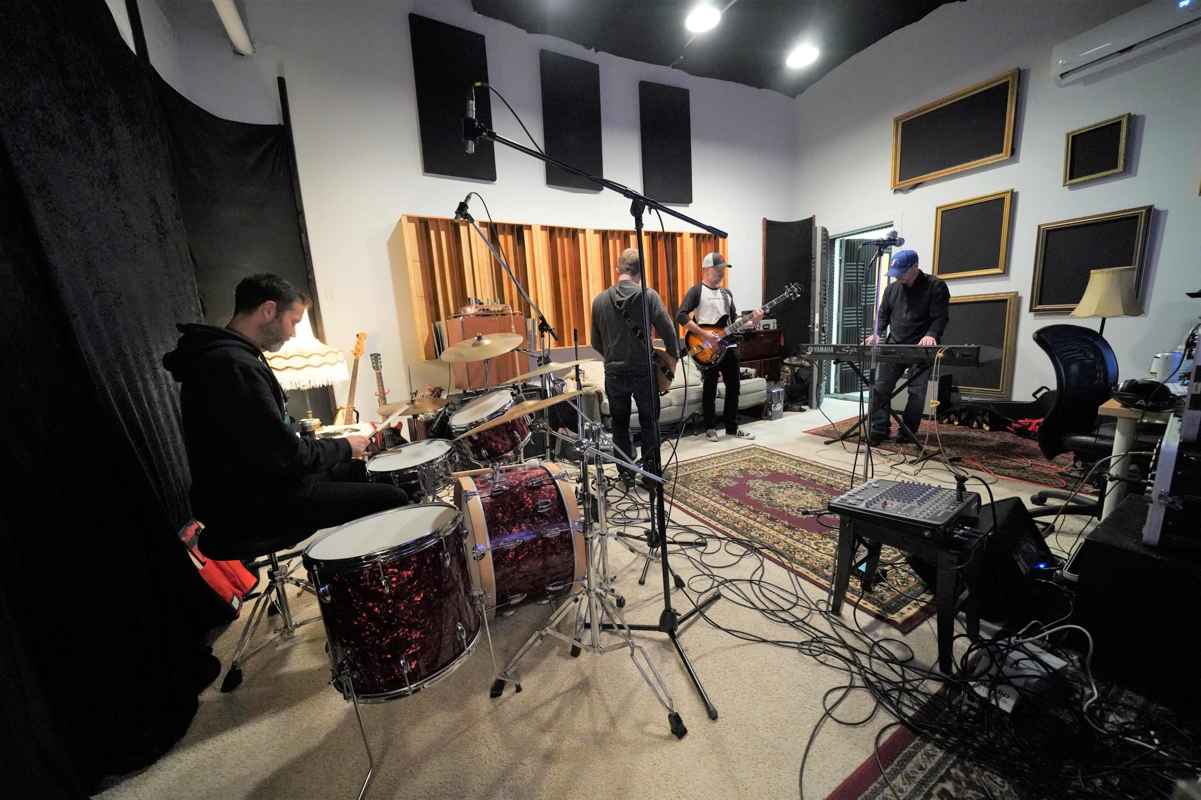 A band consisting of four musicians is rehearsing in a recording studio, with a drummer in the foreground and others playing guitar and keyboards.