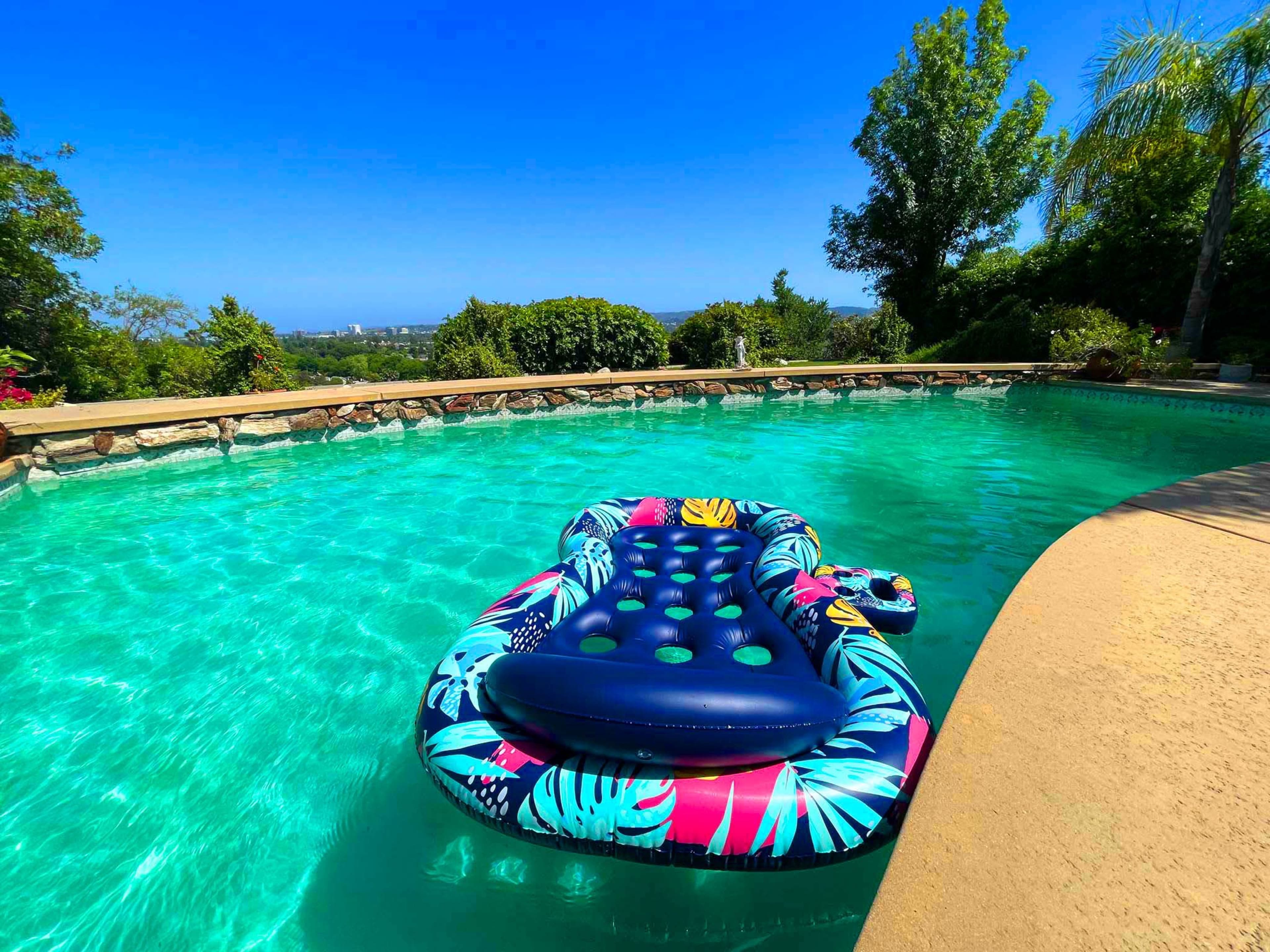 A colorful inflatable lounge floats on the surface of a clear pool surrounded by a stone deck and lush greenery.