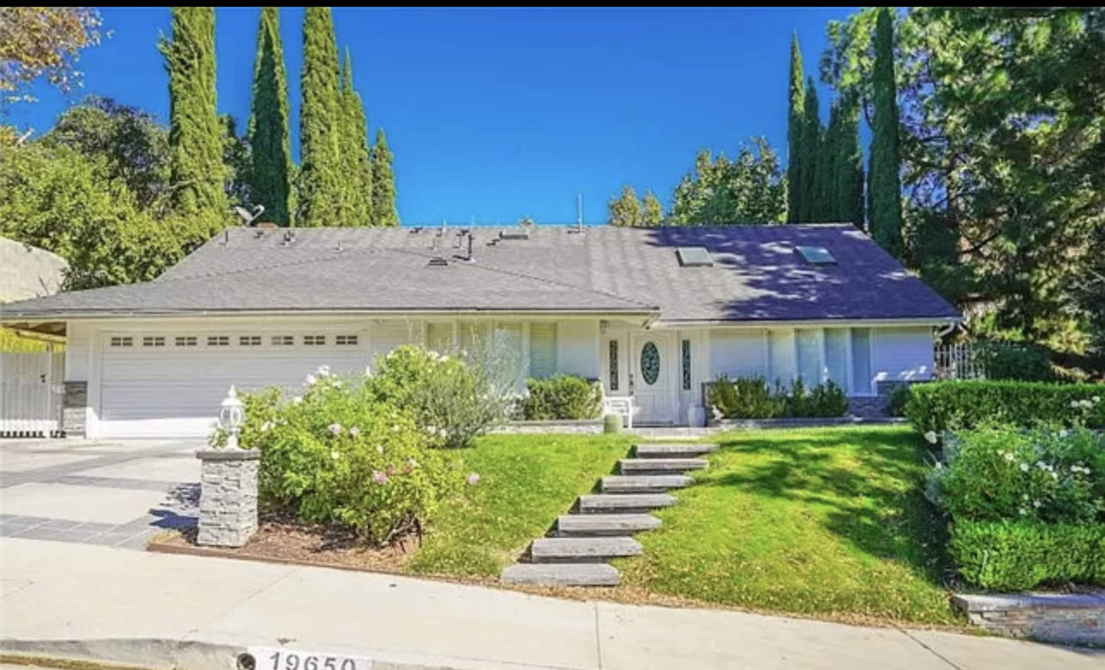 A single-story house with a gray roof, surrounded by green shrubs and tall trees, featuring a lawn and a pathway leading to the entrance.