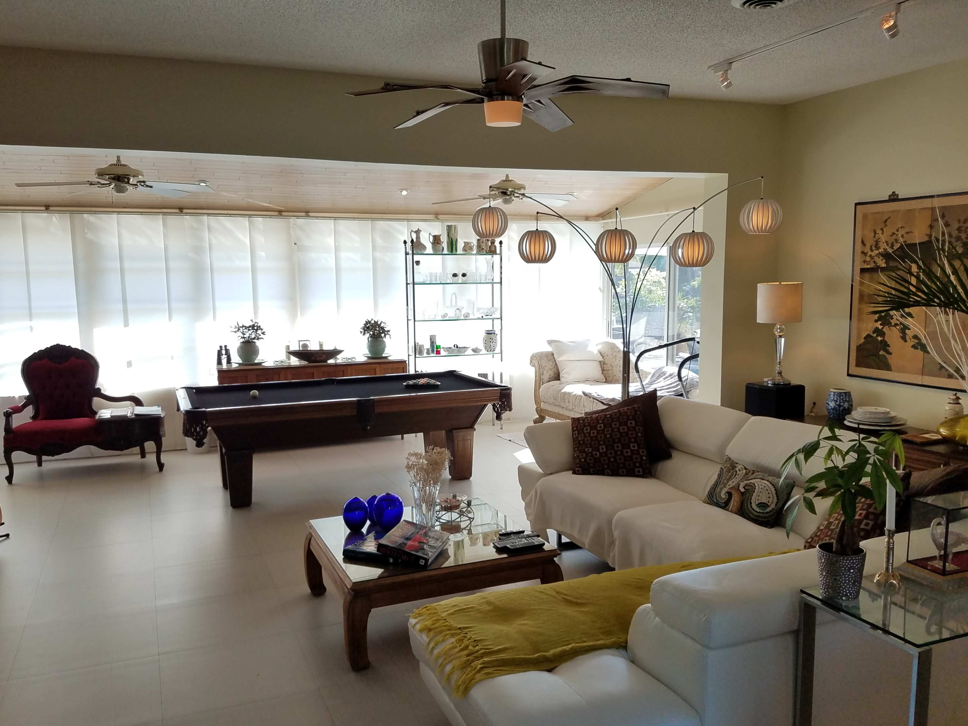 The image shows a spacious living area with a pool table, comfortable seating, decorative plants, and large windows allowing natural light to enter.