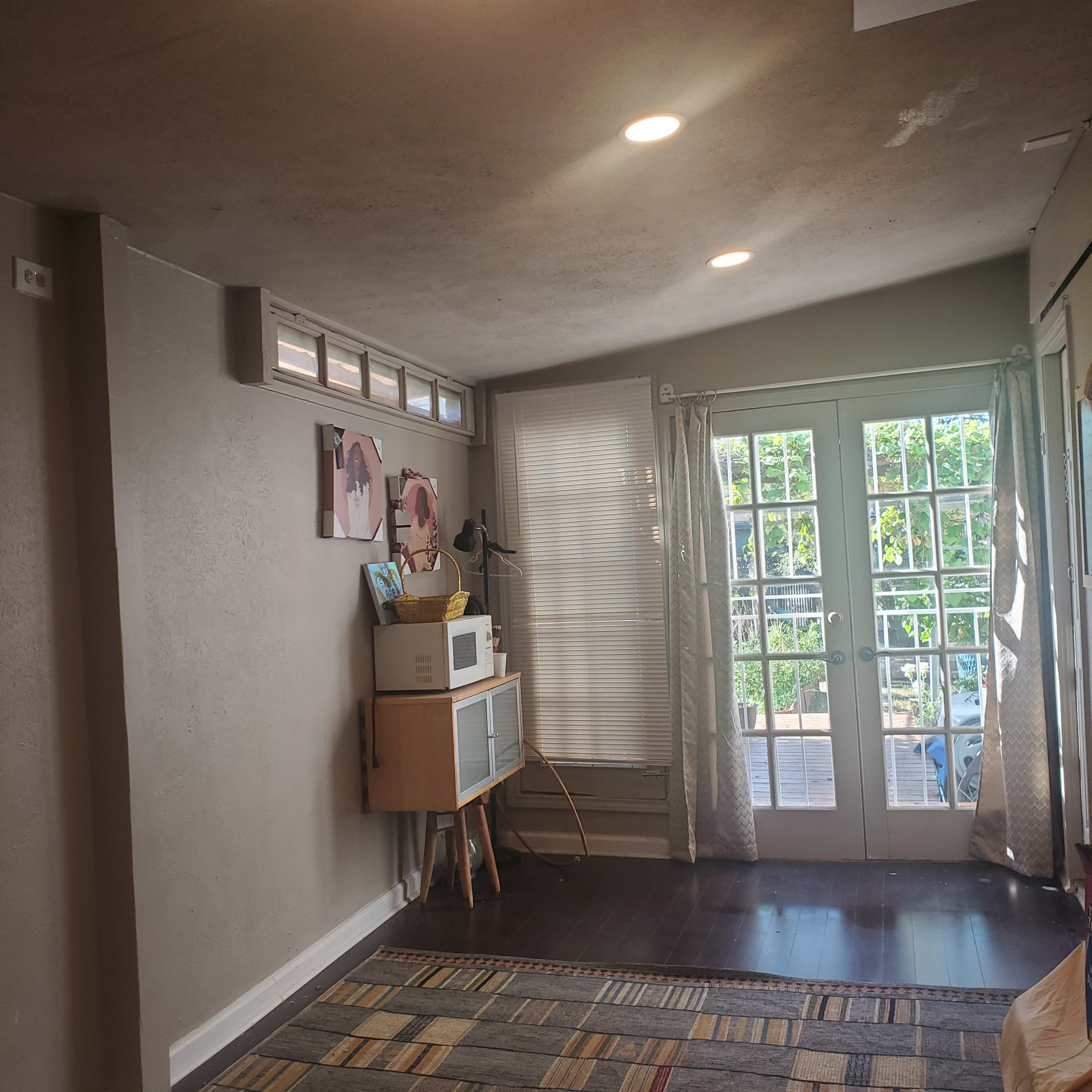 The image shows a room with a wooden floor, off-white walls, a door with multiple glass panes leading to an outdoor area, and a small cabinet with a microwave.