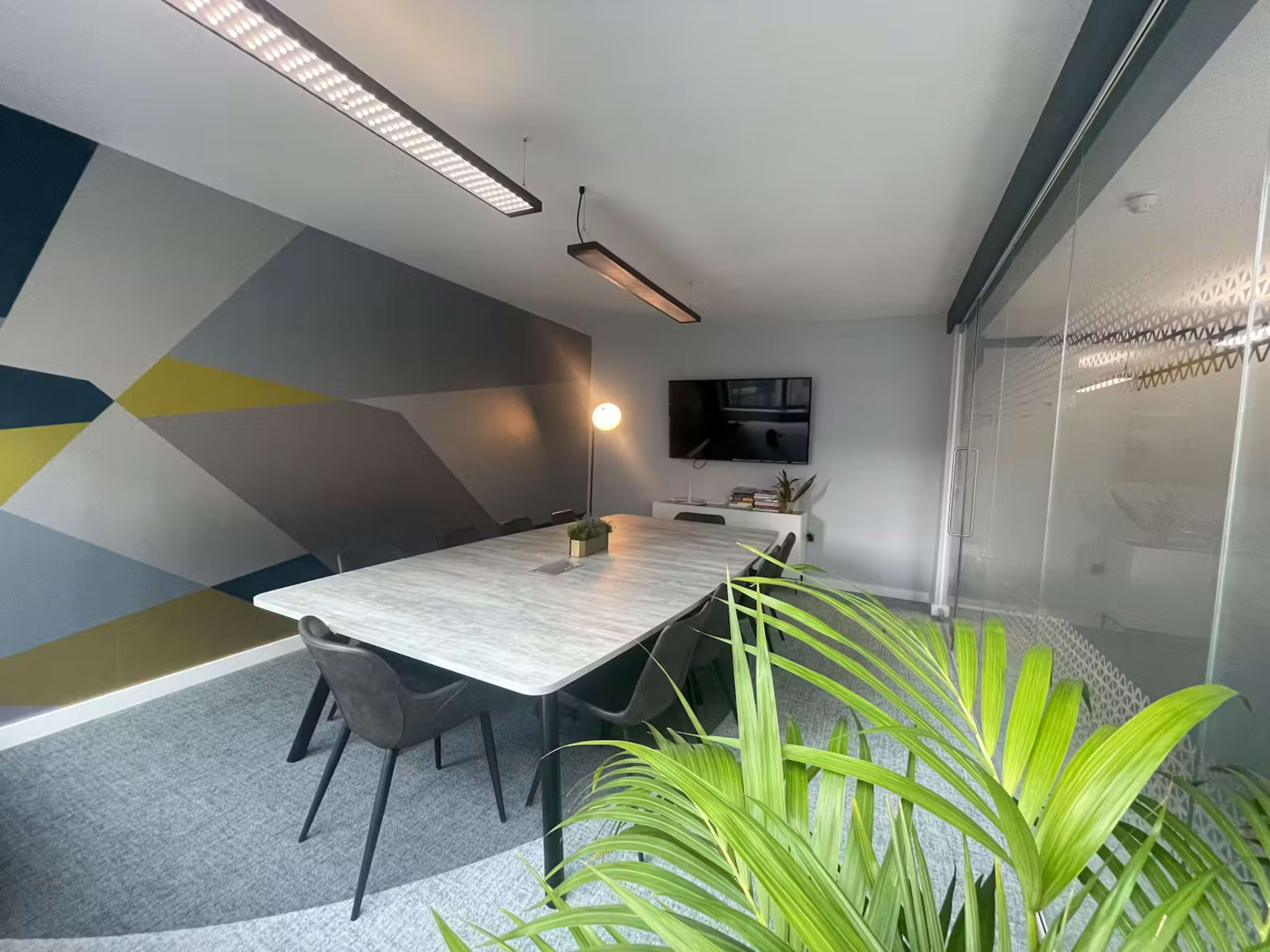 A well-lit meeting room features a large table surrounded by chairs, with a geometric wall design and a TV mounted on the wall.