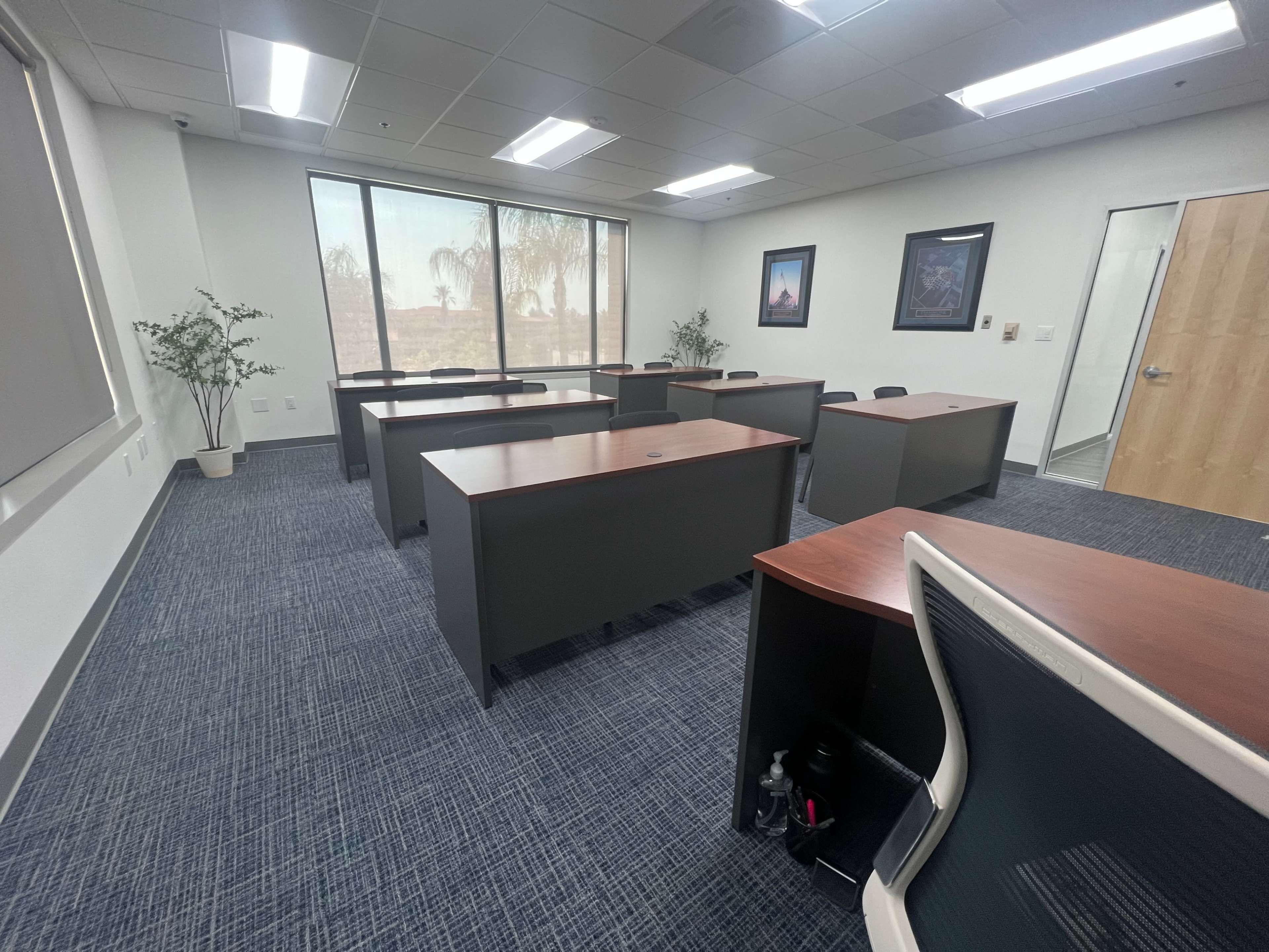 A conference room with several desks arranged in a spacious layout, a plant in one corner, and large windows letting in natural light.