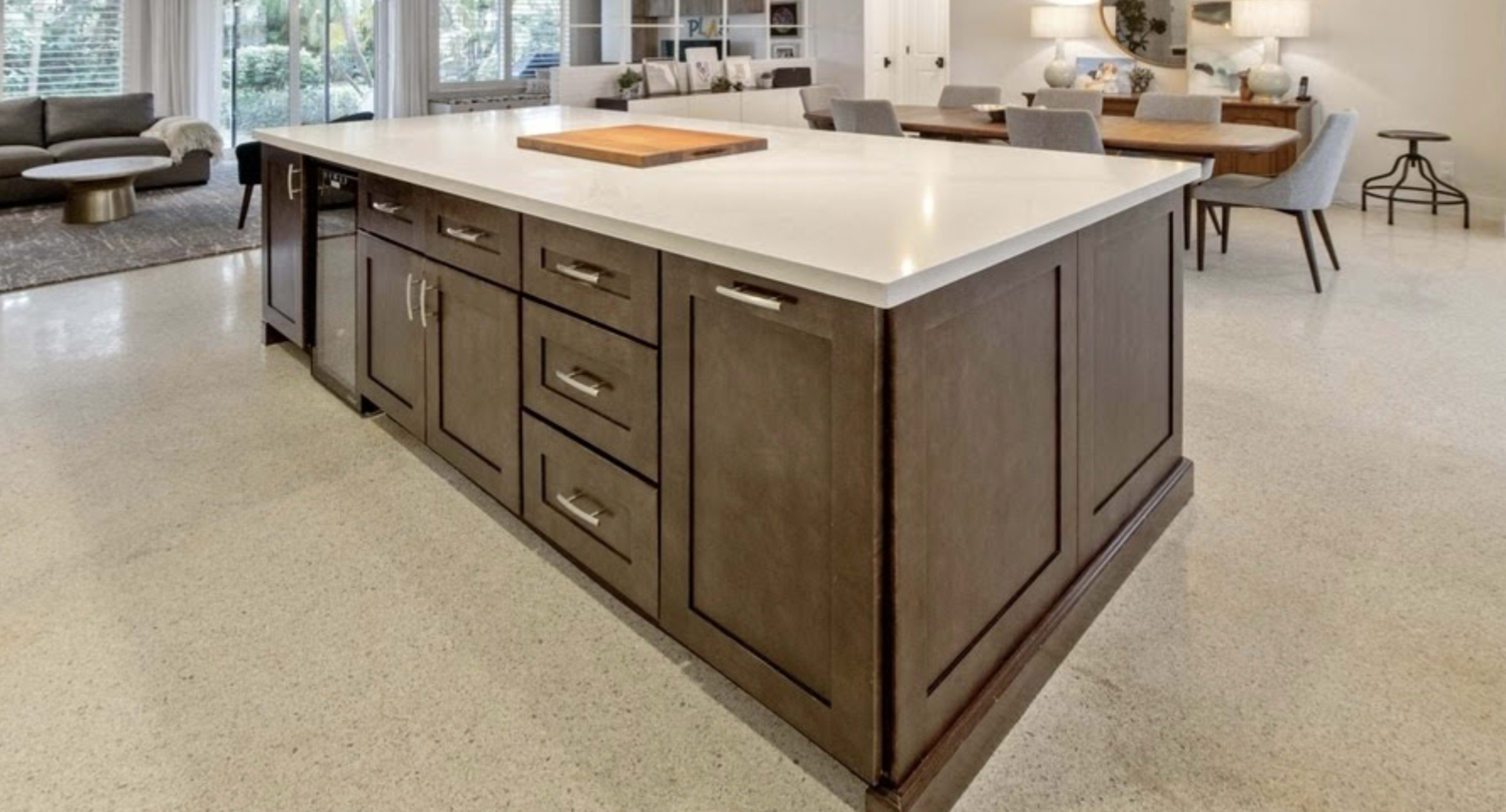 The image shows a spacious kitchen island with a light-colored countertop and dark wood cabinetry, situated near a dining area and living space.