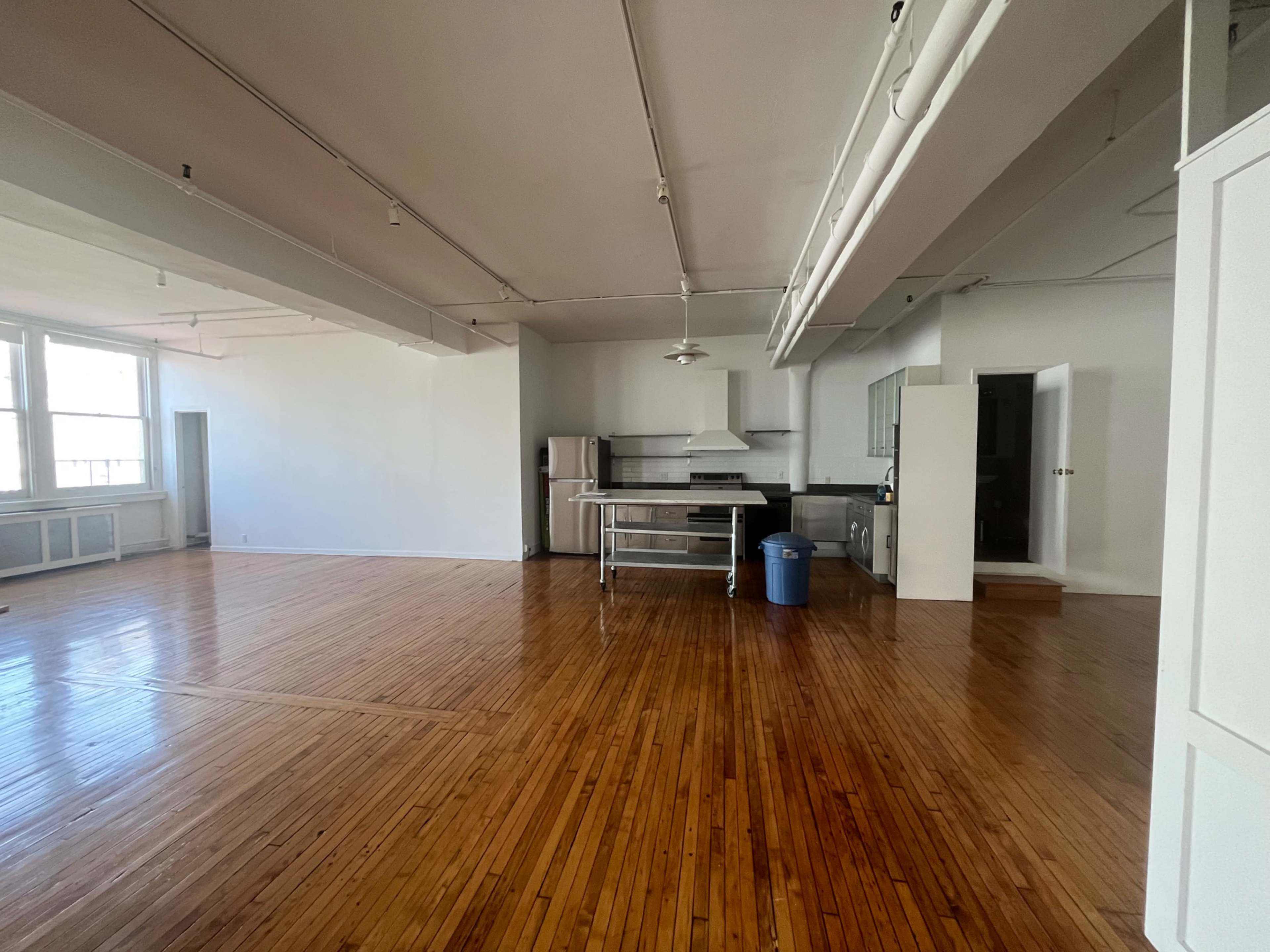 The image shows a spacious, empty room with polished hardwood floors, a kitchen area along one wall, and large windows allowing natural light.