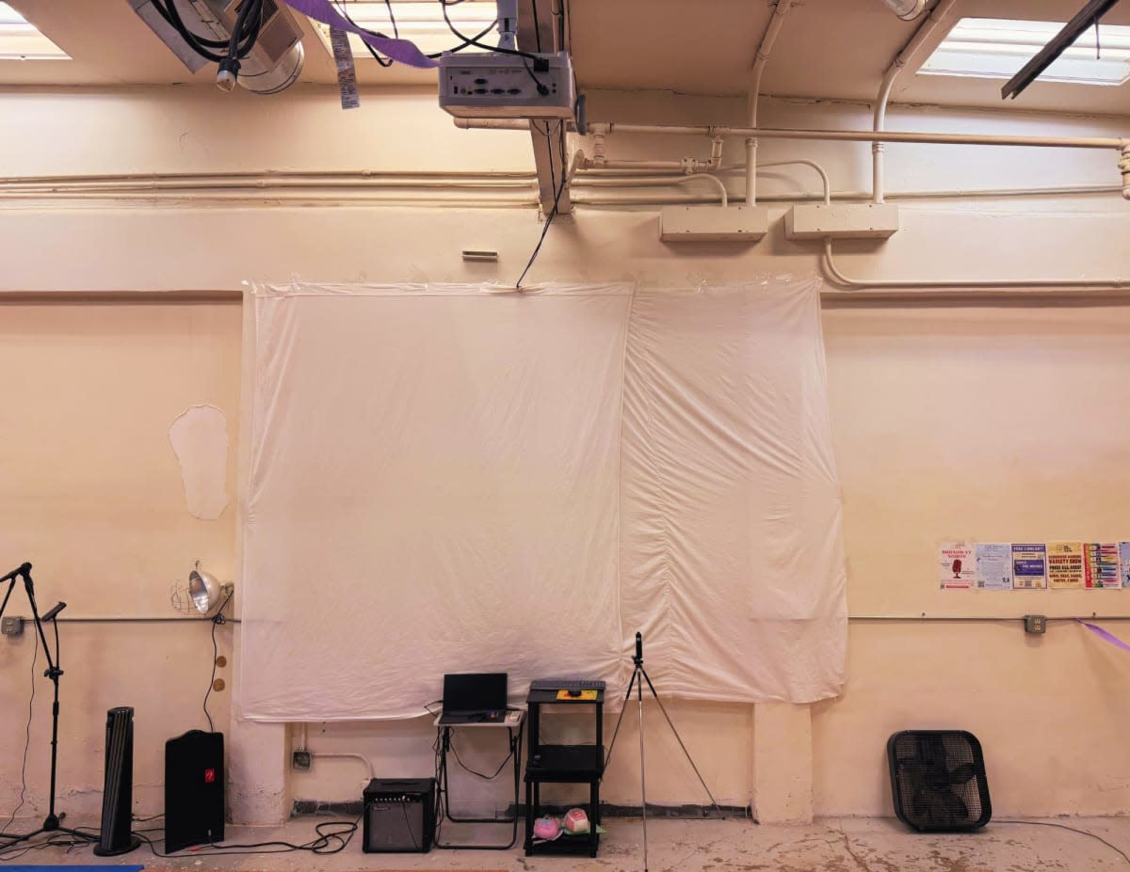 The image shows a simple room with two large white sheets hanging on a beige wall, a laptop on a small black cart, and a speaker nearby.