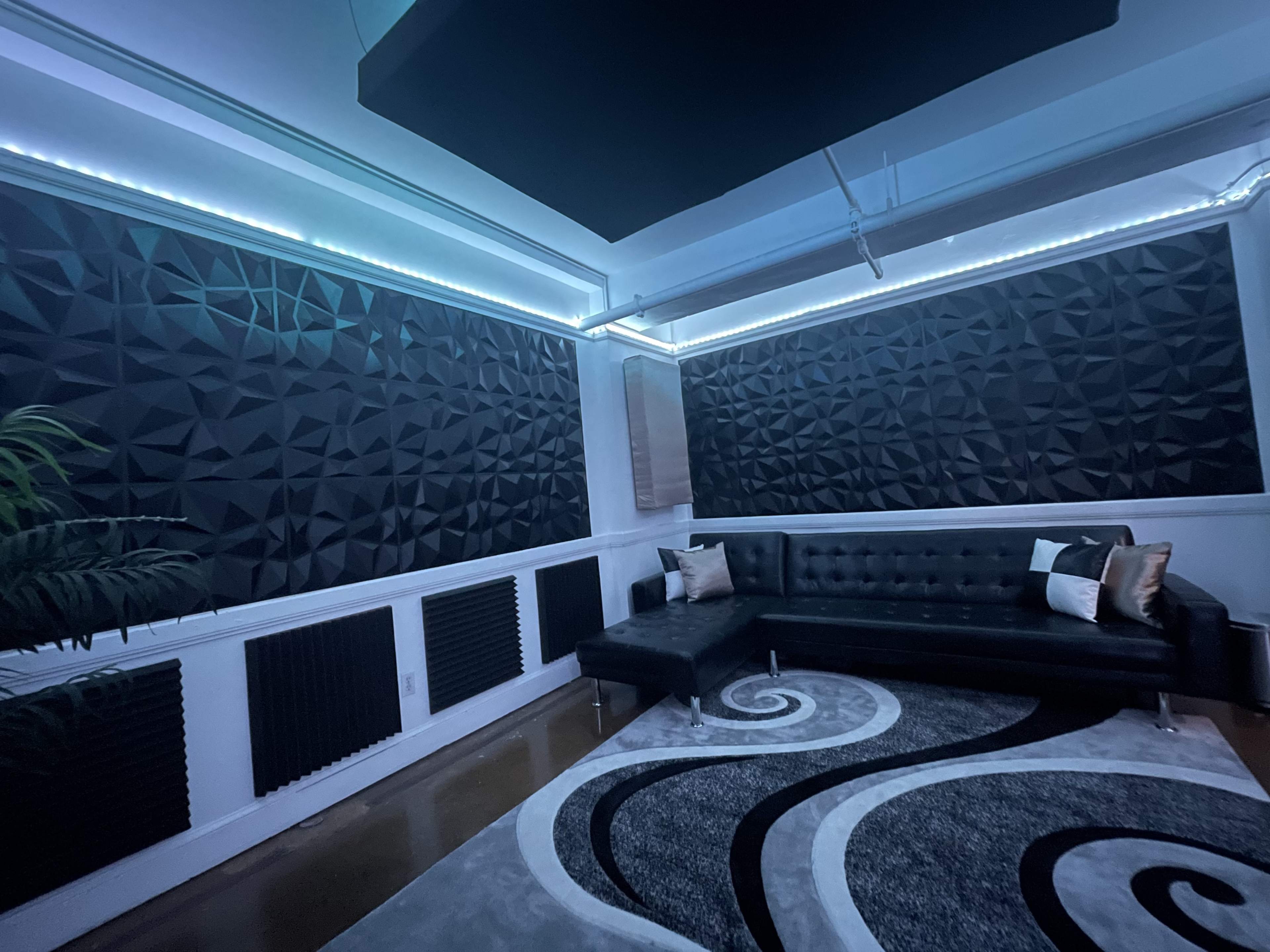 A modern room with a black leather sectional couch, angular acoustic wall panels, and a patterned area rug.