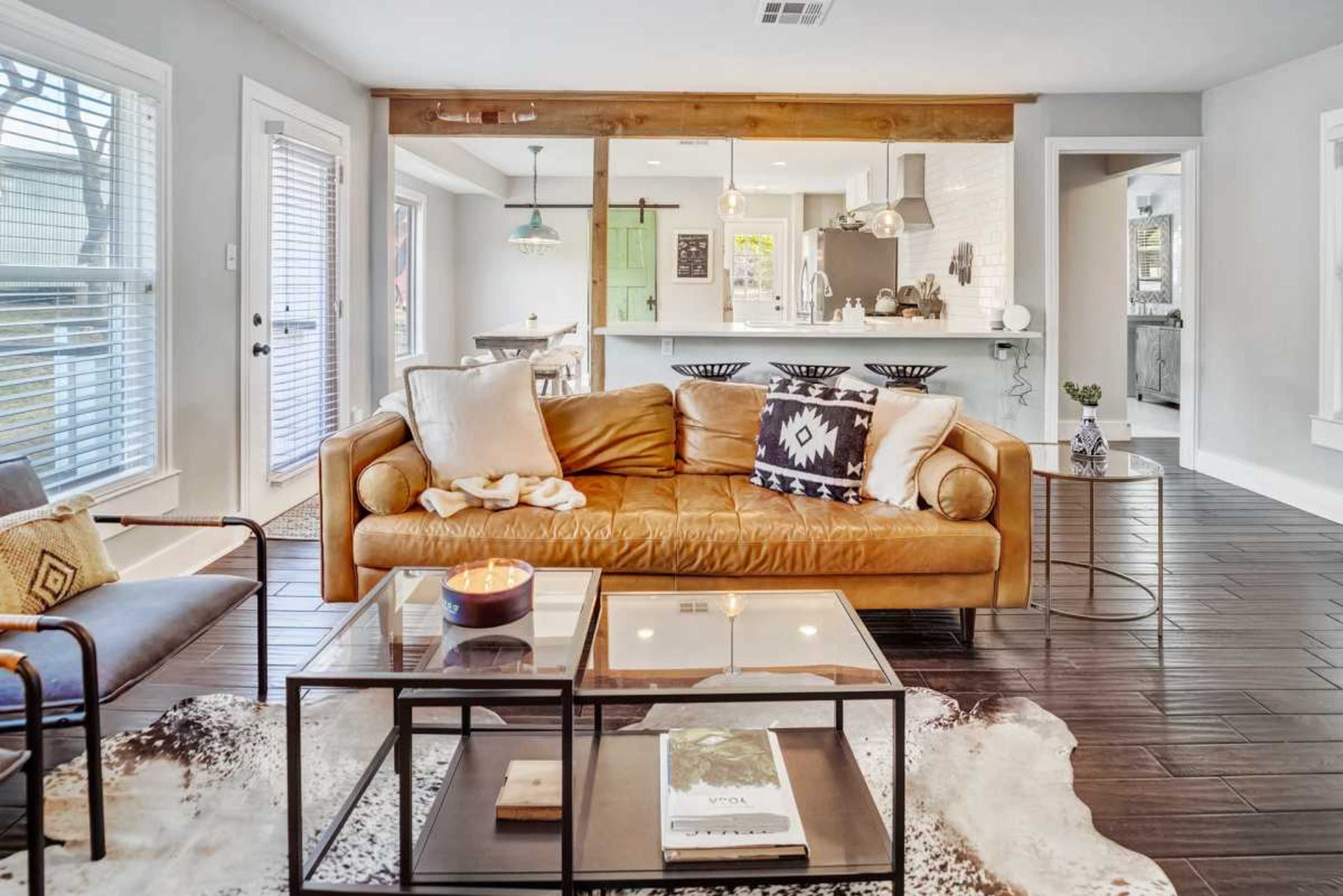 The image shows a stylish living room with a leather sofa, a glass coffee table, and a view into a modern kitchen area featuring light-colored cabinets and a dining space.