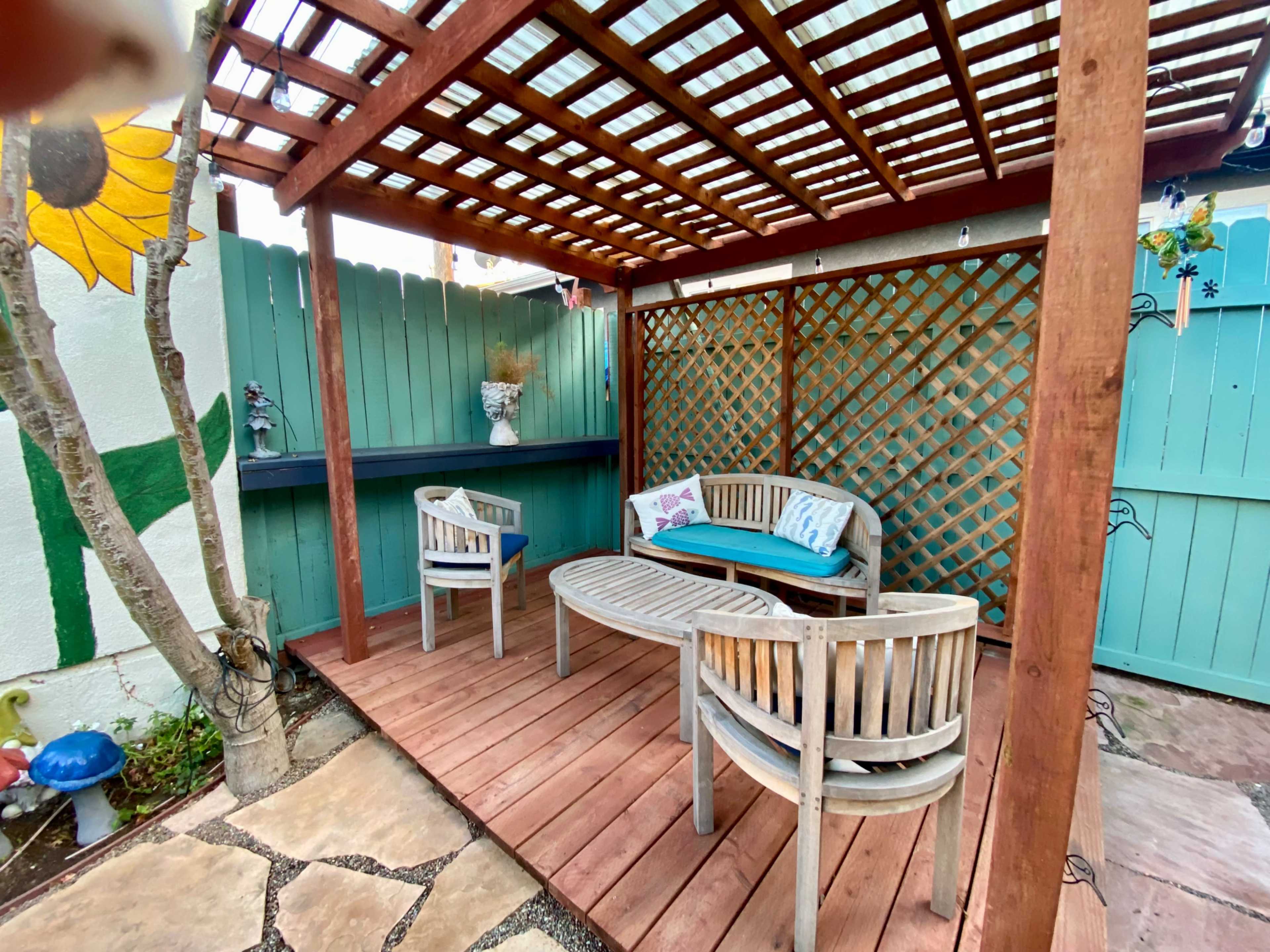 A wooden patio area with a lattice roof, featuring a circular wooden table and three chairs, with a painted sunflower and a turquoise fence in the background.