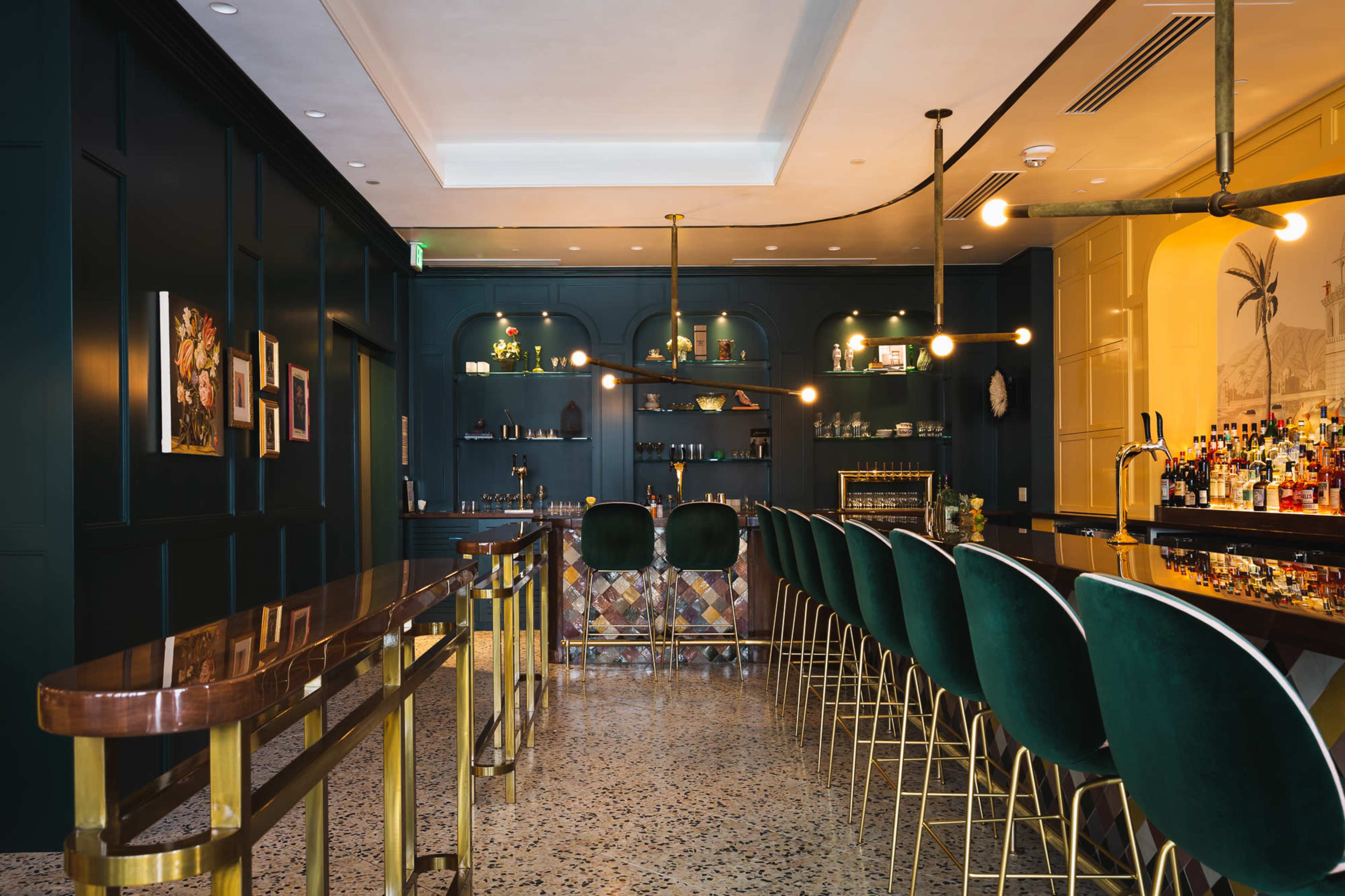 The image shows a stylish bar area with a dark green wall, sleek bar stools, and a well-stocked shelf of bottles.