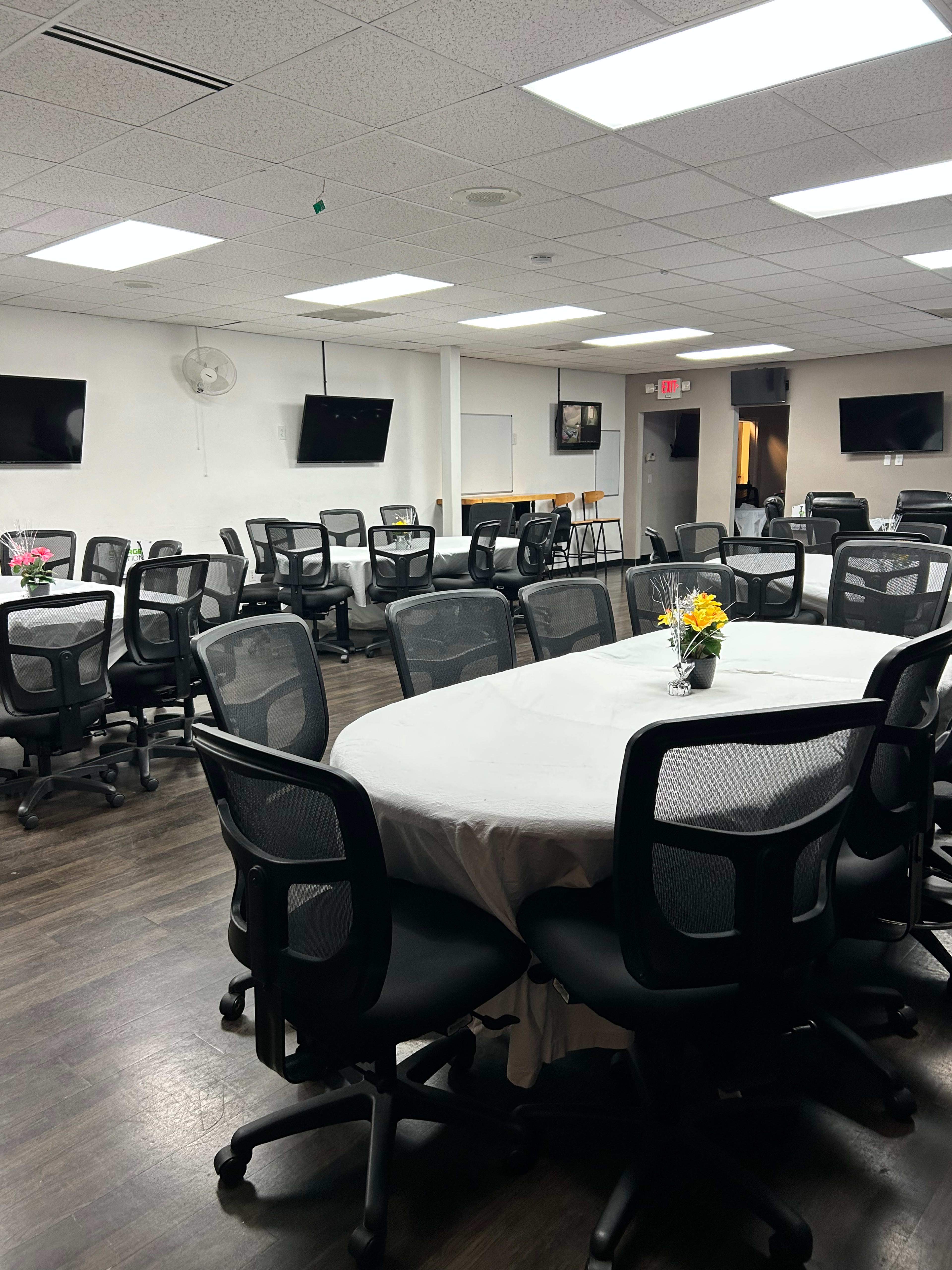 A conference room with multiple circular tables, each covered with a tablecloth, surrounded by ergonomic office chairs and equipped with mounted televisions on the walls.