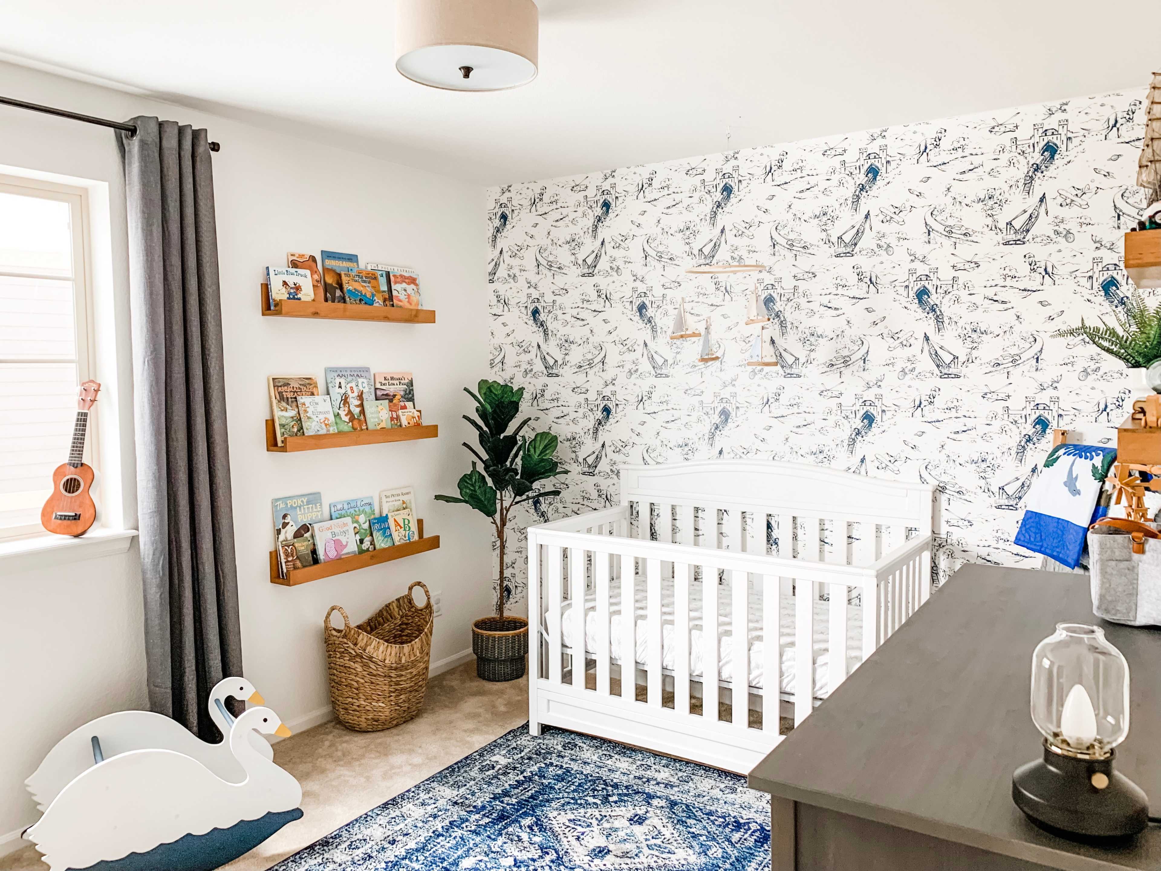 The room features a white crib, bookshelves filled with children's books, a potted plant, and a blue rug, all set against a patterned wallpaper.