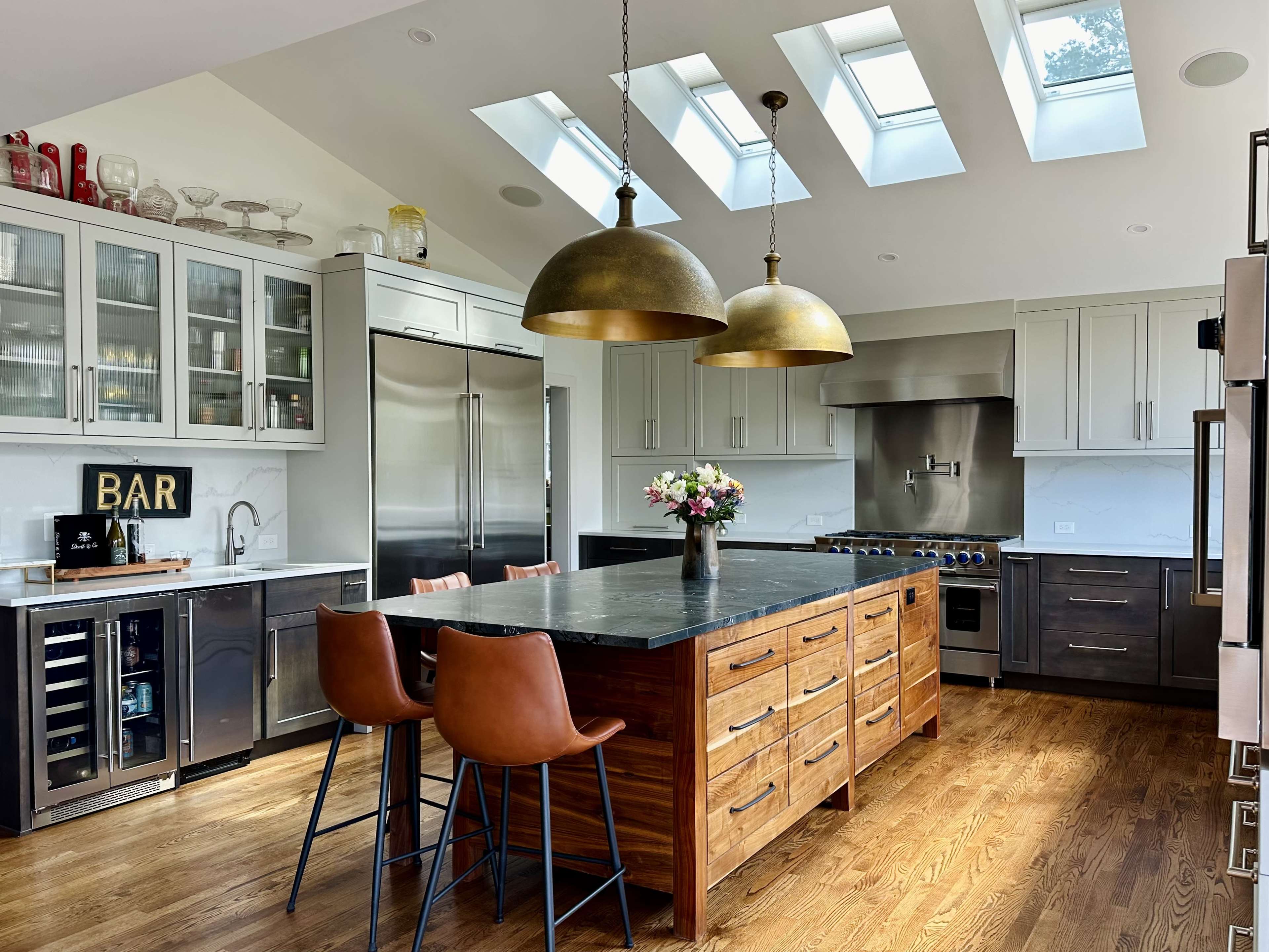 The kitchen features a central wooden island with four chairs, stainless steel appliances, glass-front cabinets, and skylights above.