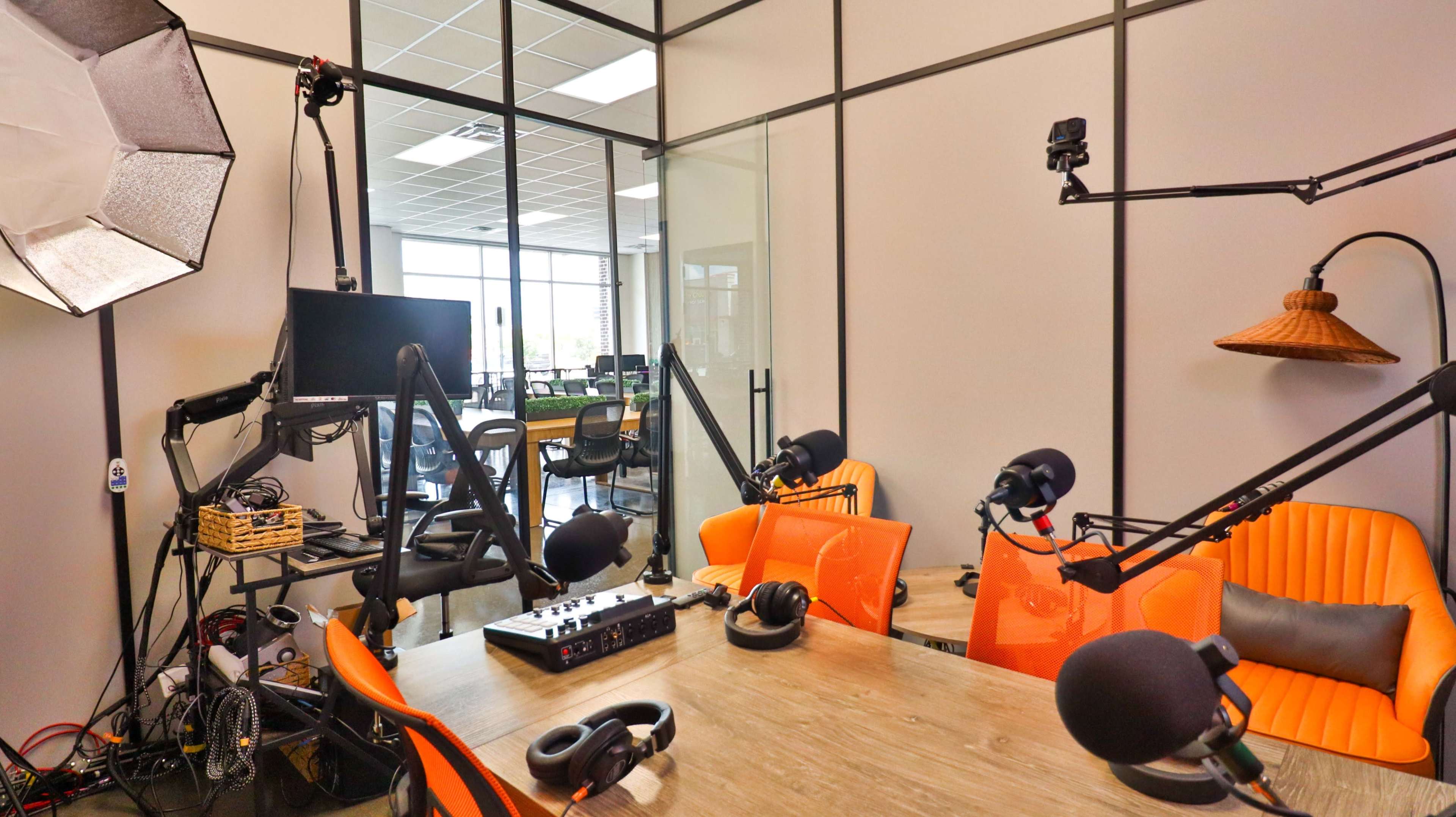The image shows a modern podcast recording space featuring a wooden table with orange chairs, microphones on stands, and video equipment, all enclosed in a glass-walled studio.