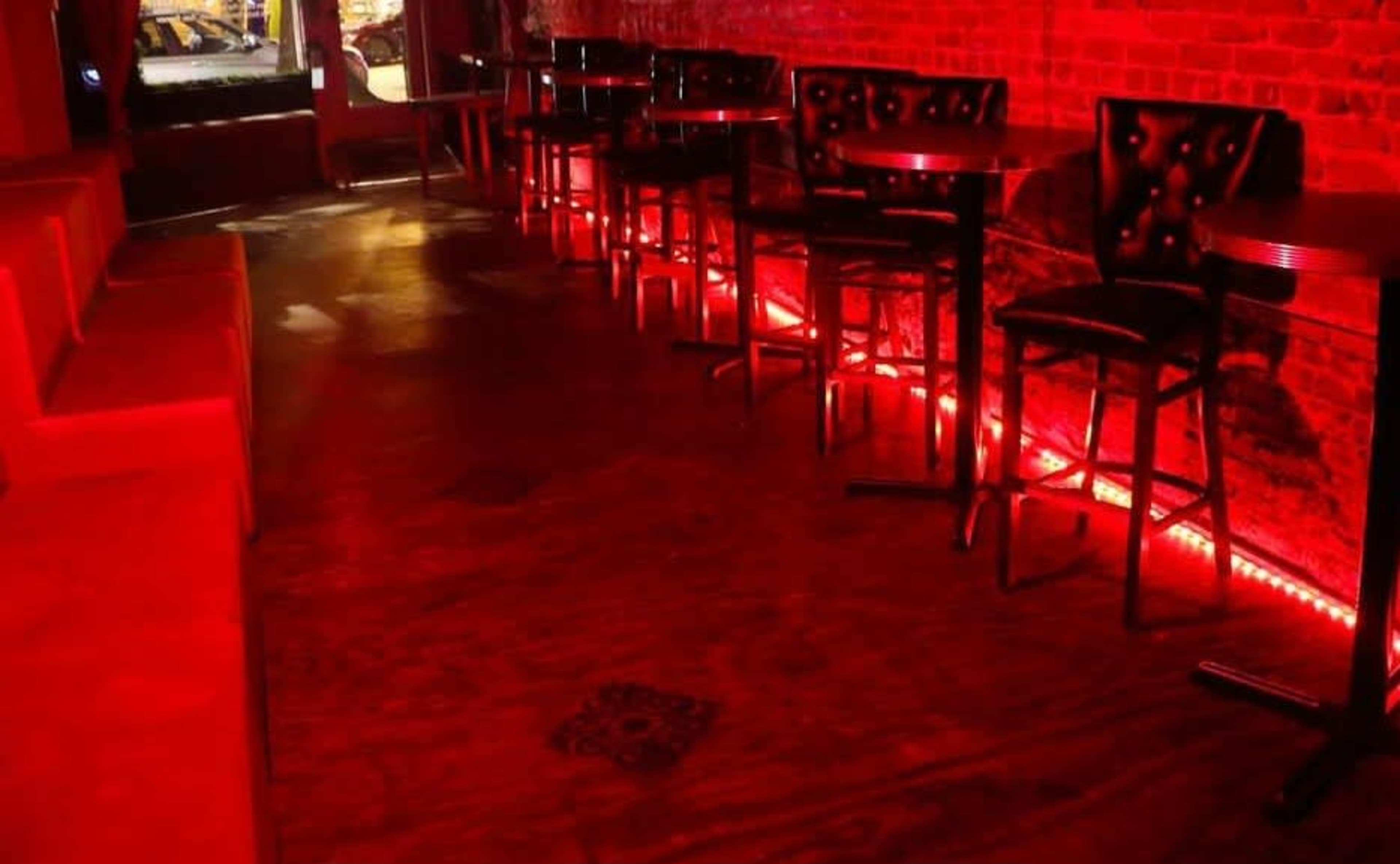 The image shows a dimly lit bar interior with red lighting, featuring wooden flooring and several tall tables with black chairs along a brick wall.