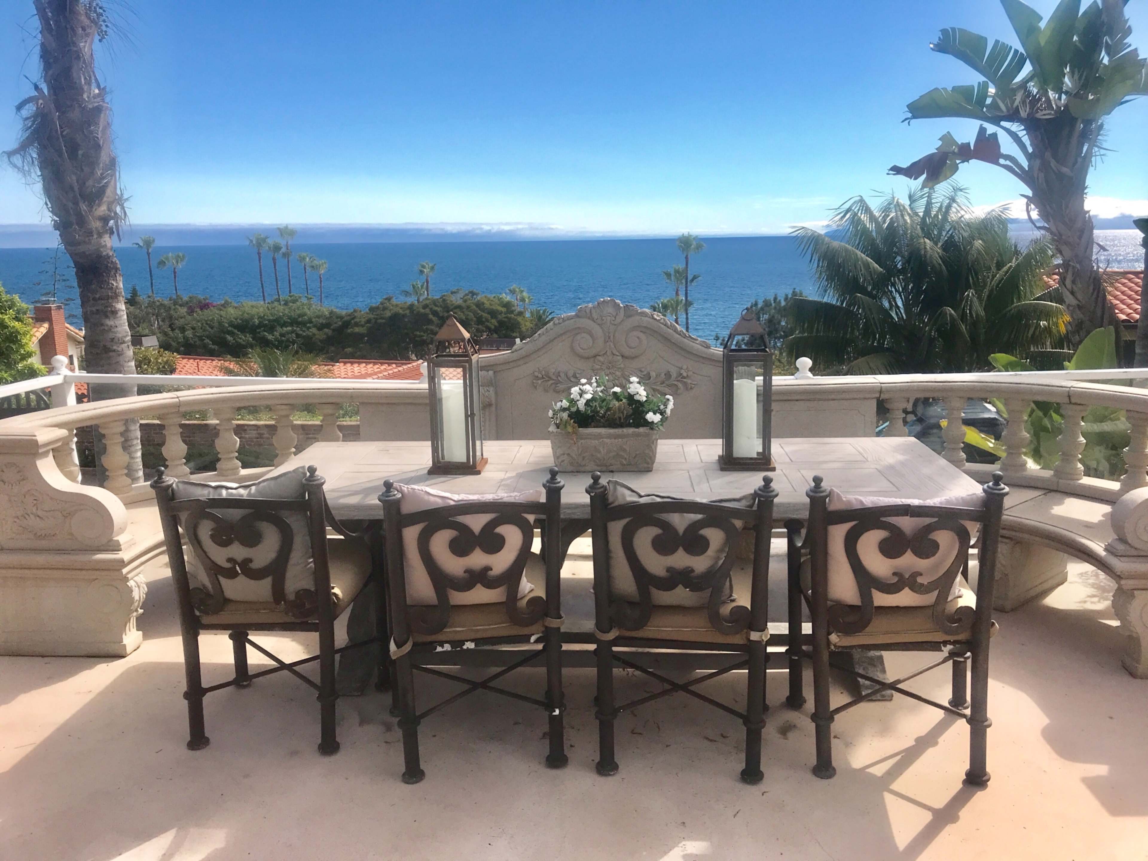A wooden dining table with ornate metal chairs stands on a terrace overlooking the ocean.