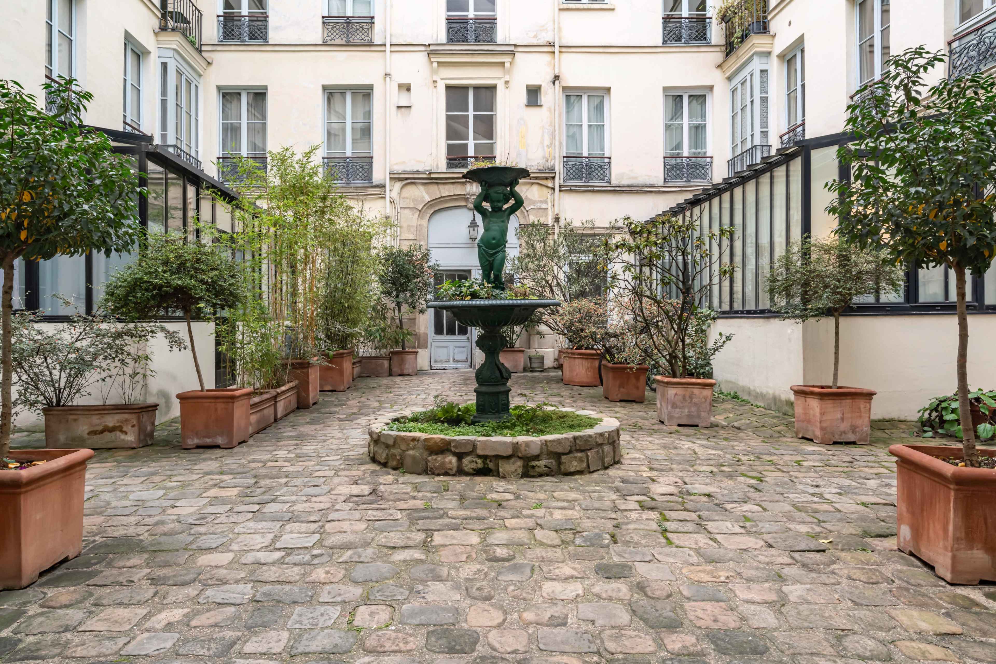The image depicts a courtyard with a central green fountain surrounded by potted plants and stone pavement.