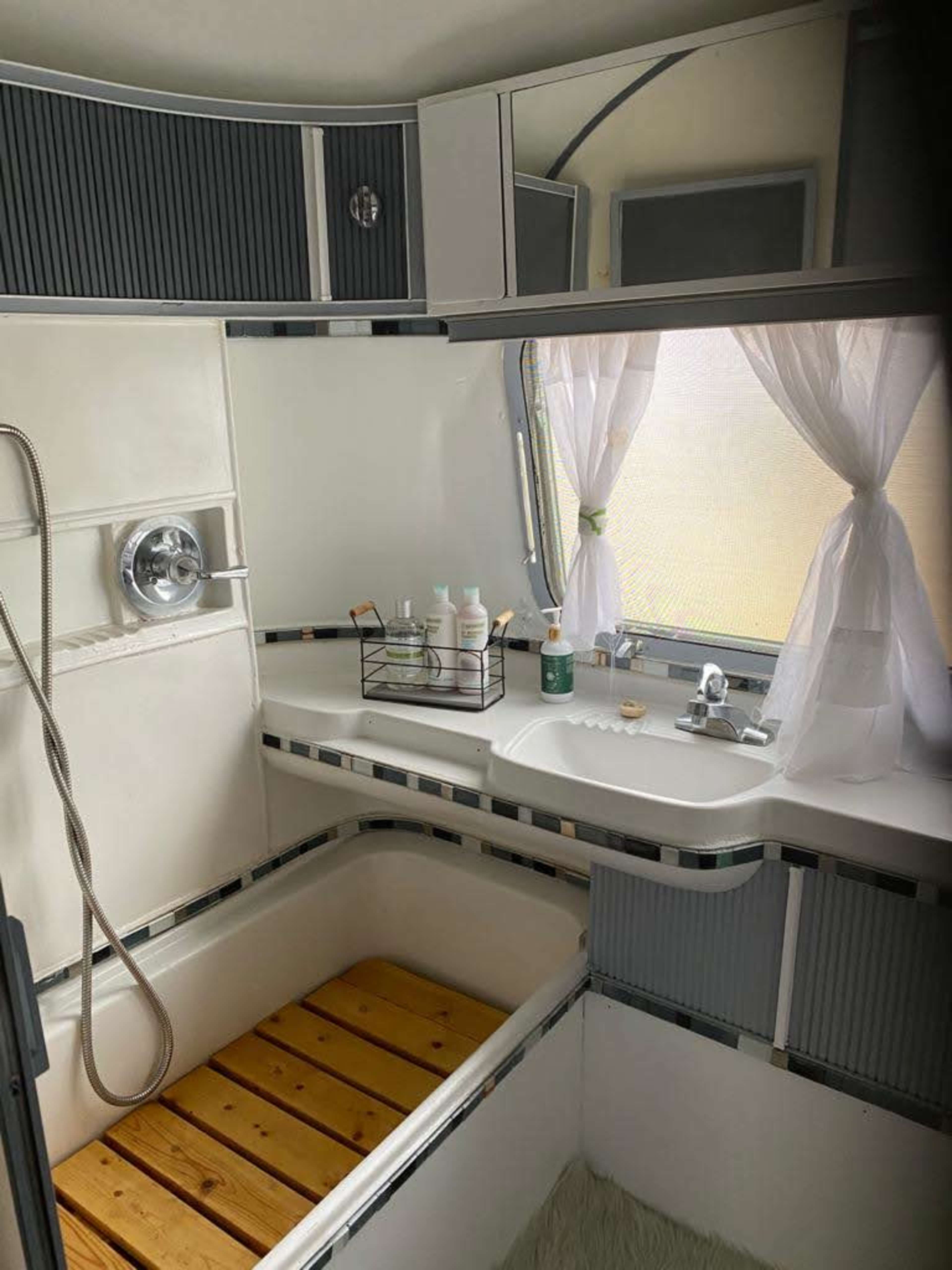 The image shows a compact bathroom featuring a wooden slatted bathtub, sink area with various toiletries, and a showerhead attached to the wall.