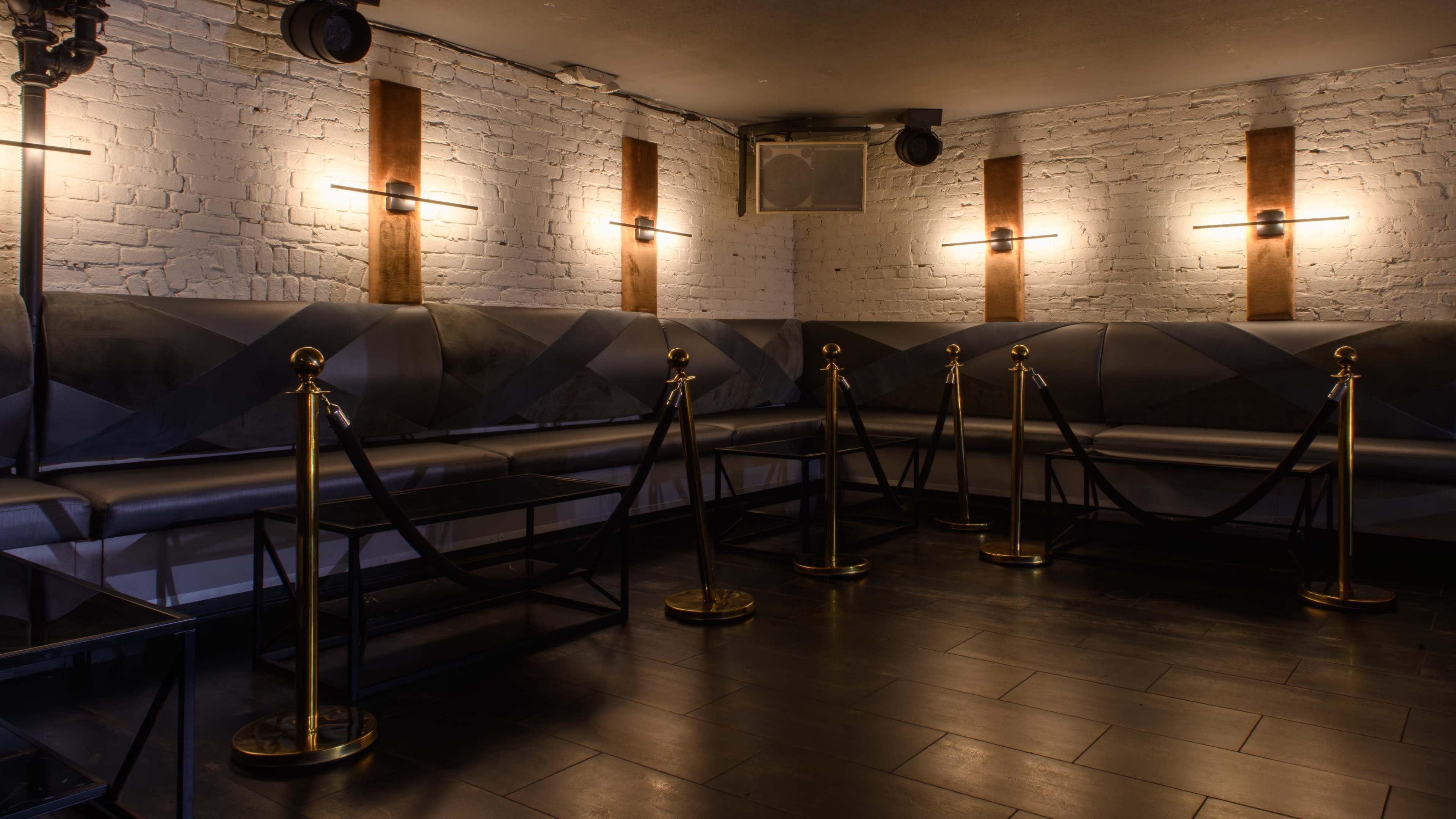 The image shows a dimly lit lounge area with gray seating, wooden accents, and gold stanchions separating the seating from the walkway.