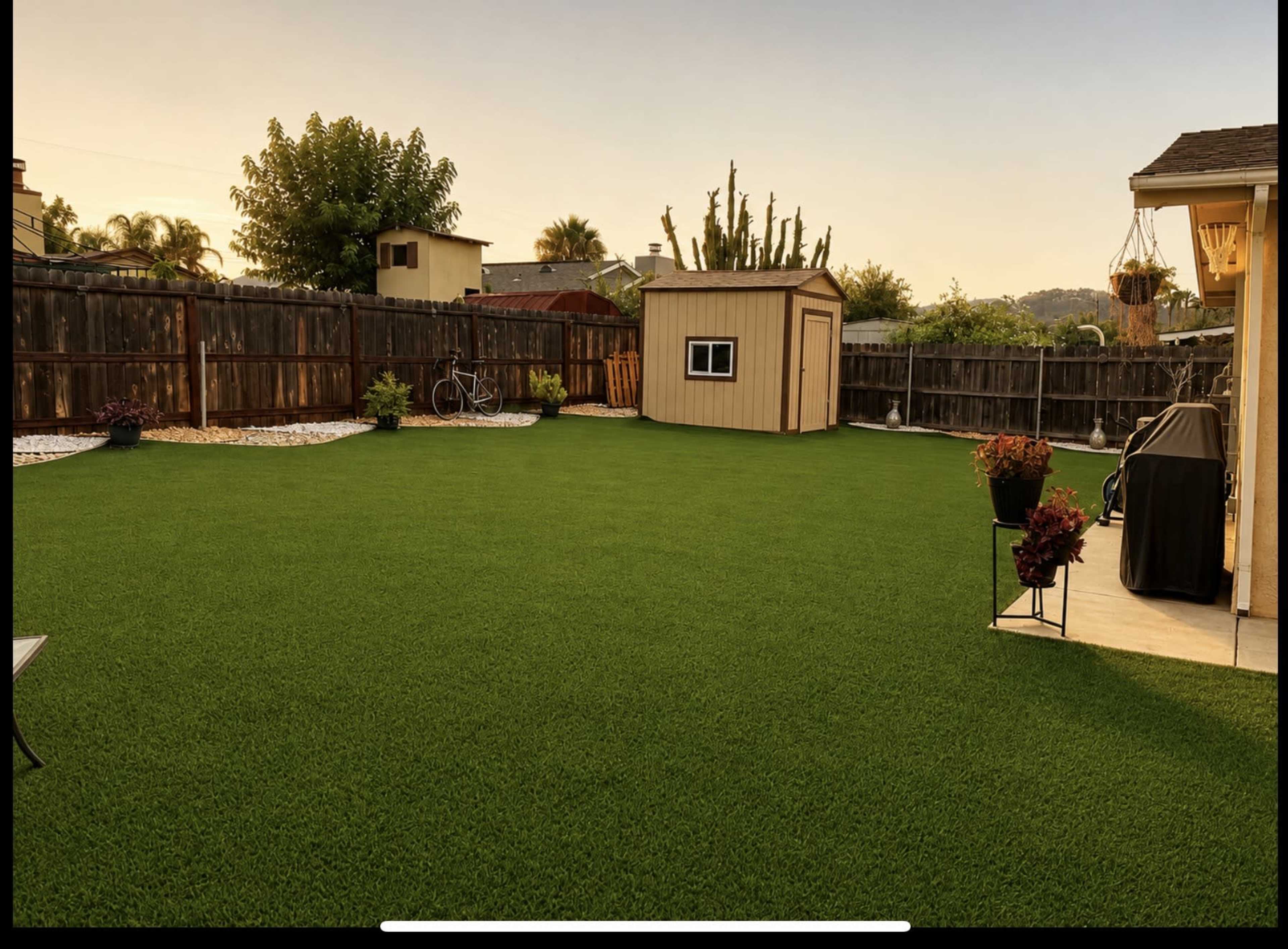 The image shows a spacious backyard with artificial grass, a storage shed, a barbecue grill, and several potted plants, surrounded by a wooden fence.