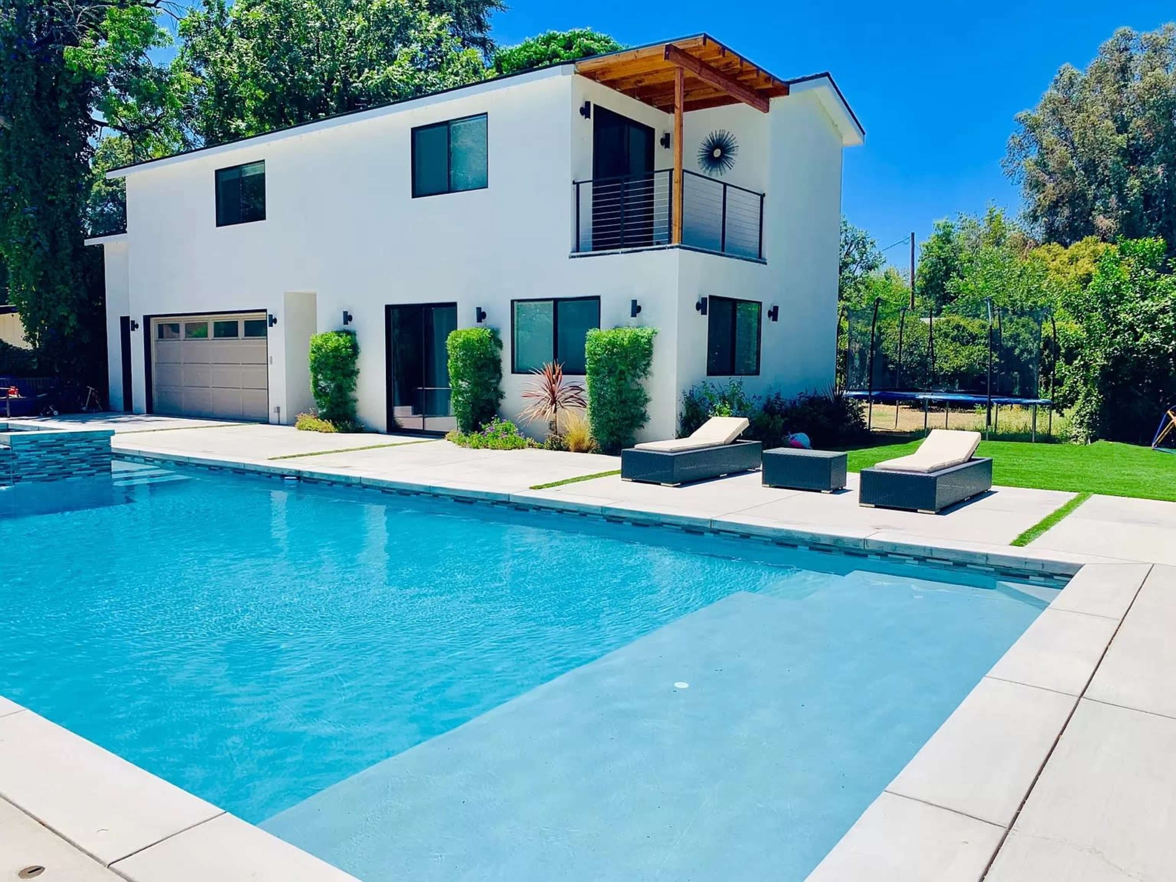 The image shows a modern two-story house with a swimming pool and lounge chairs on a sunny day.