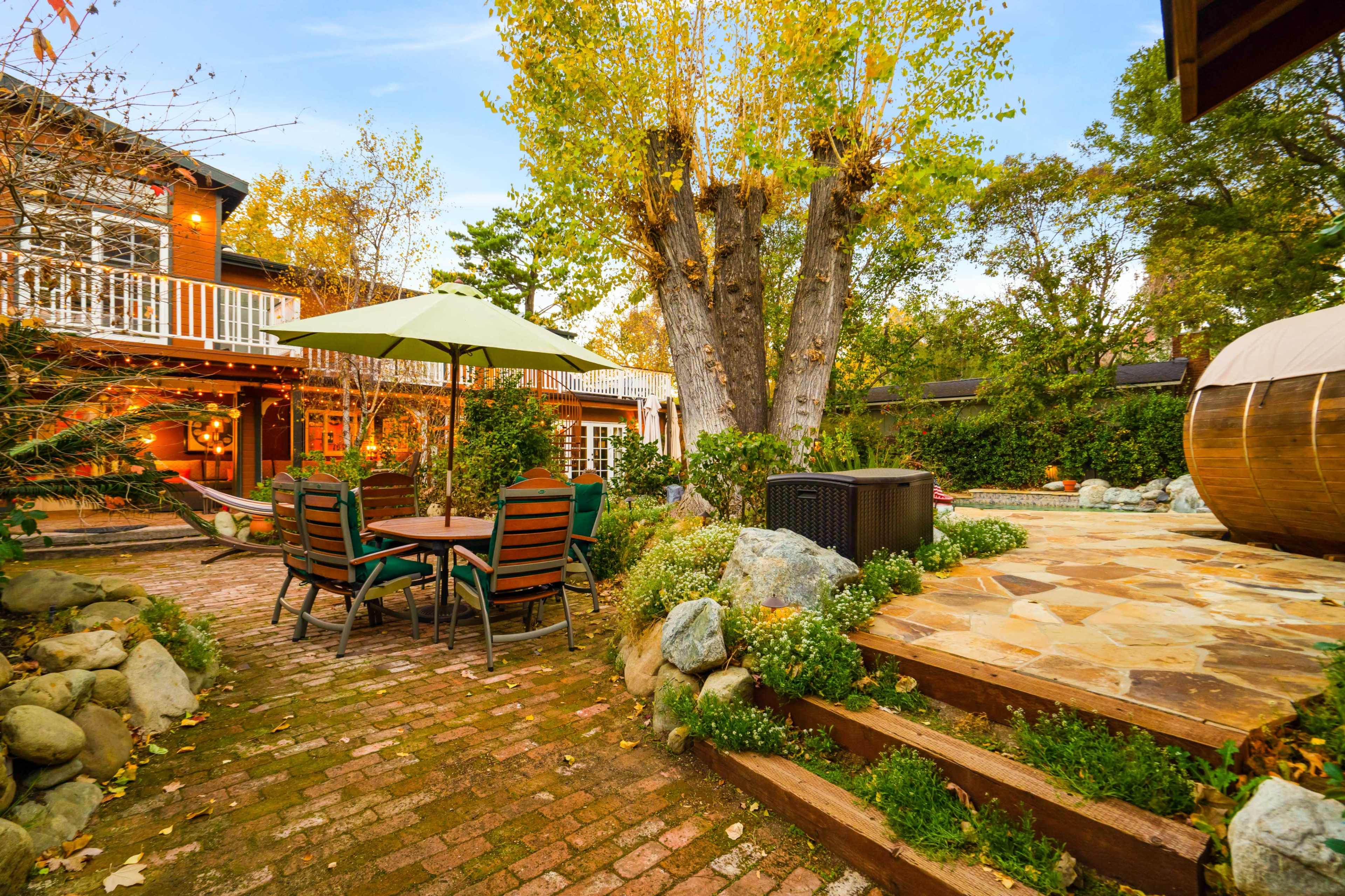 The image shows a brick patio area with a table and chairs, a green umbrella, a large tree in the background, and a wooden structure nearby.