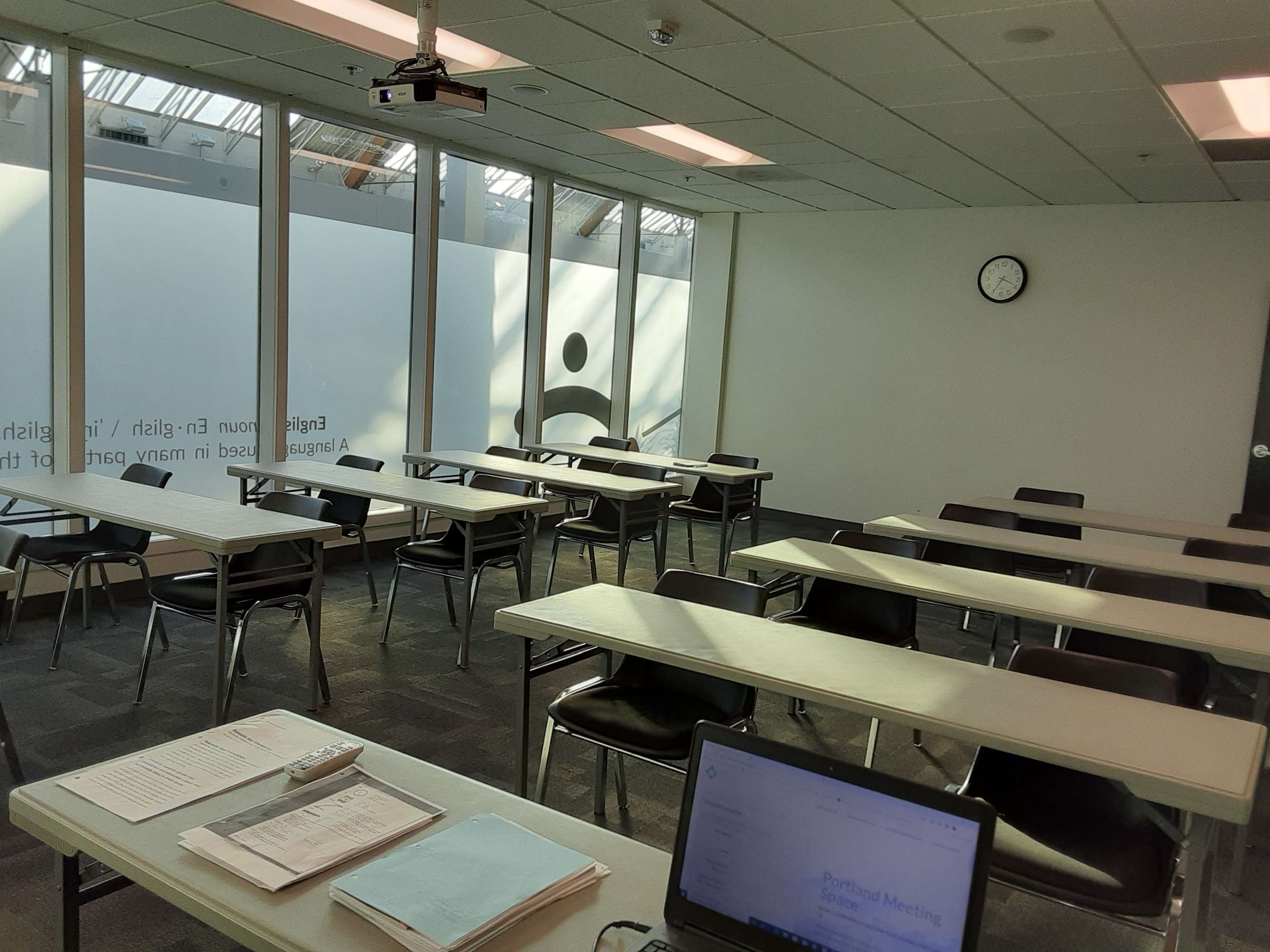 A meeting room is arranged with several rows of tables and chairs, featuring a wall clock and a laptop on the front desk.