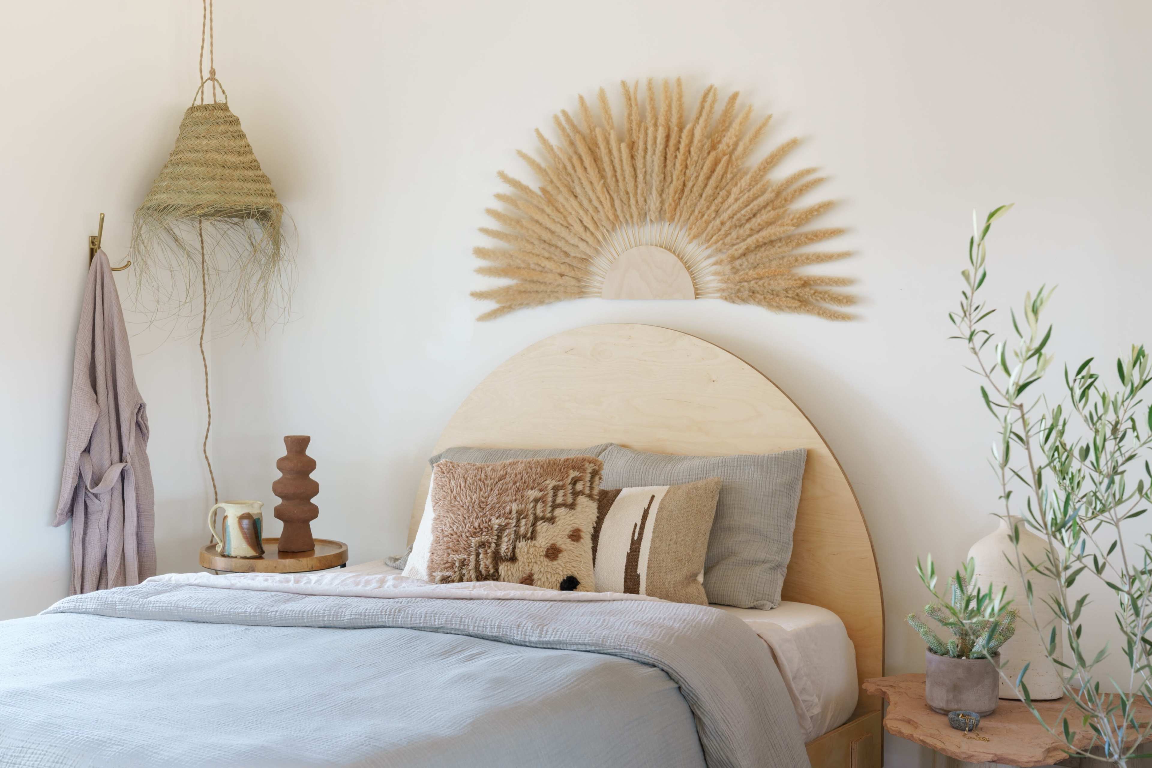 A bedroom features a wooden headboard with a decorative fan-shaped dried grass arrangement above, complemented by a bedside table with a vase and a textured throw on the bed.