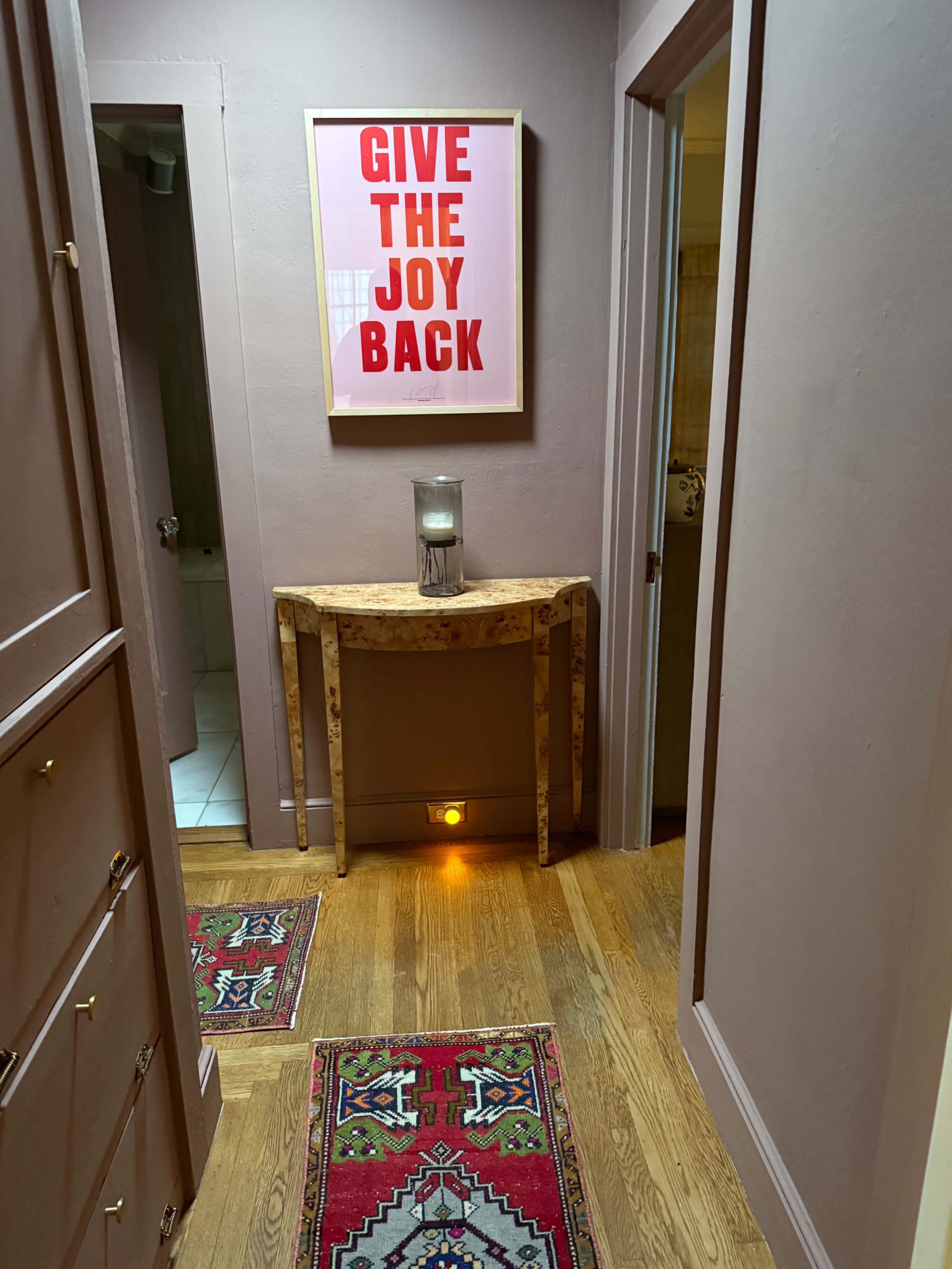 A narrow hallway features a framed pink poster with bold red text, a wooden console table, and patterned rugs on the floor.