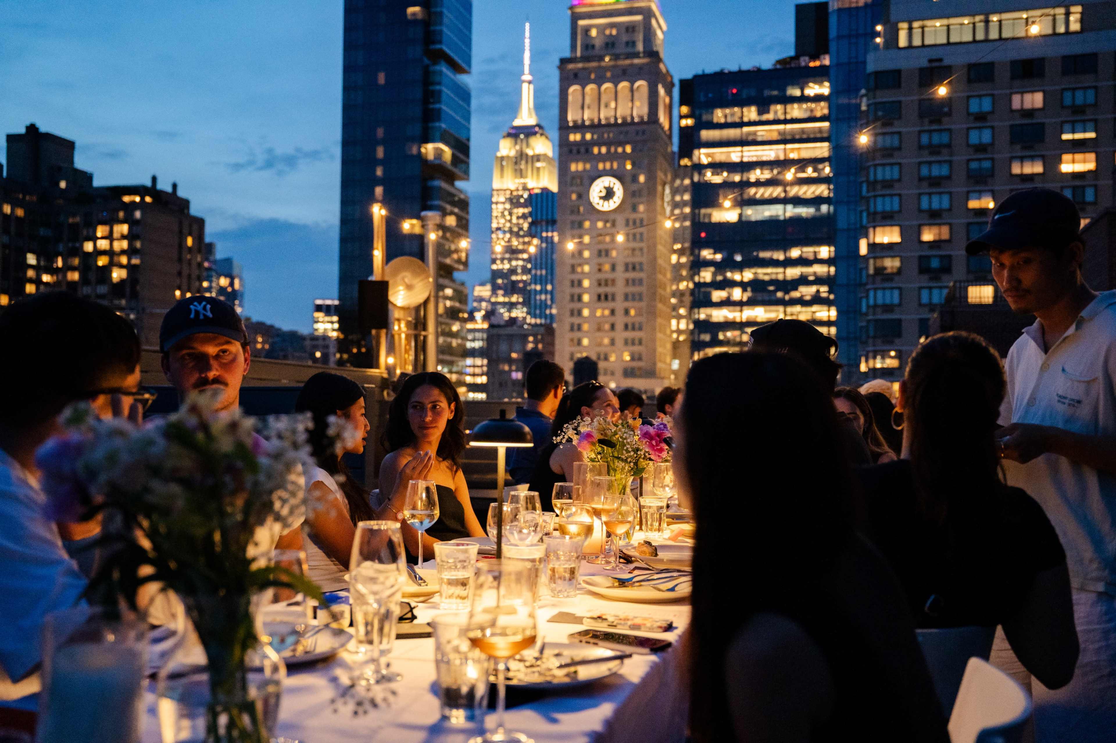A group of people is dining at a long table in an outdoor setting with the Empire State Building visible in the background, surrounded by city skyscrapers at dusk.