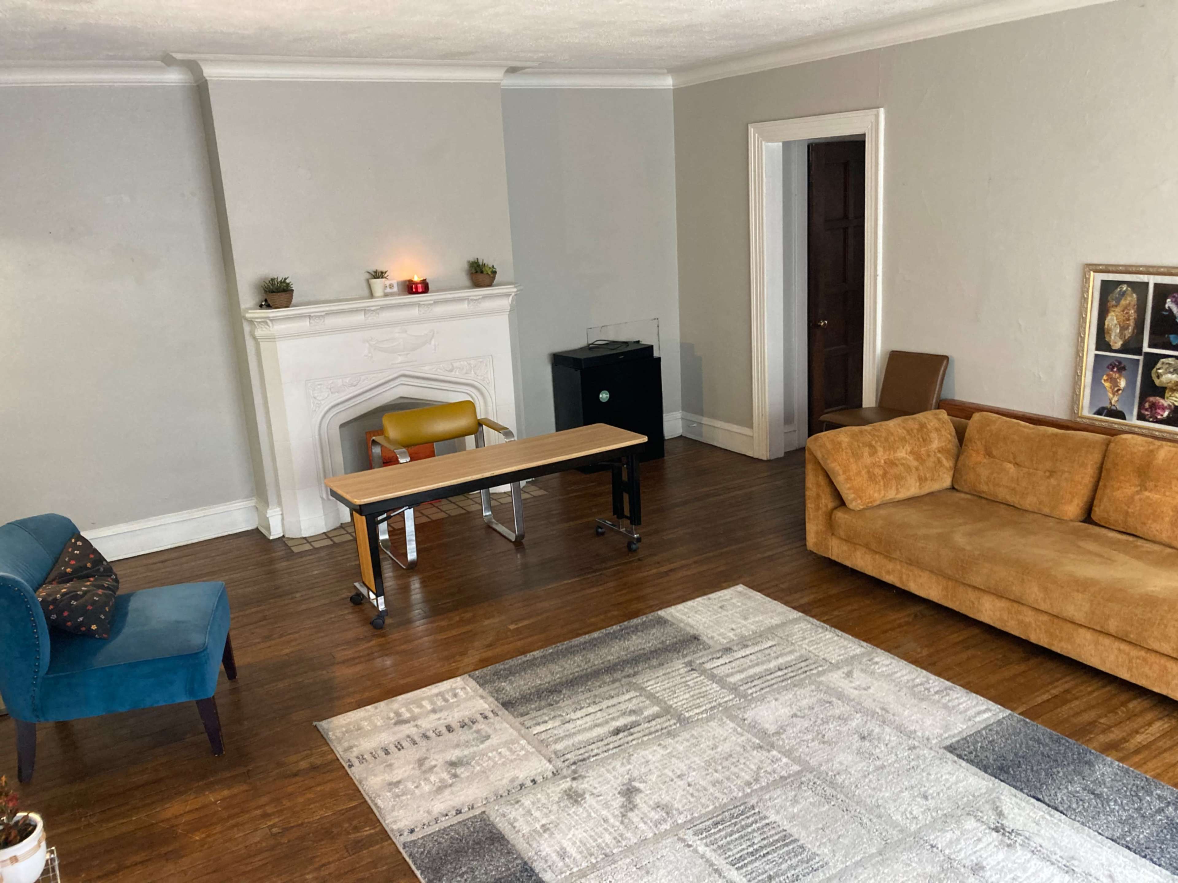 The room features a brown sofa, a blue armchair, a wooden table on wheels, a fireplace with small plants, and a large patterned rug on the wooden floor.