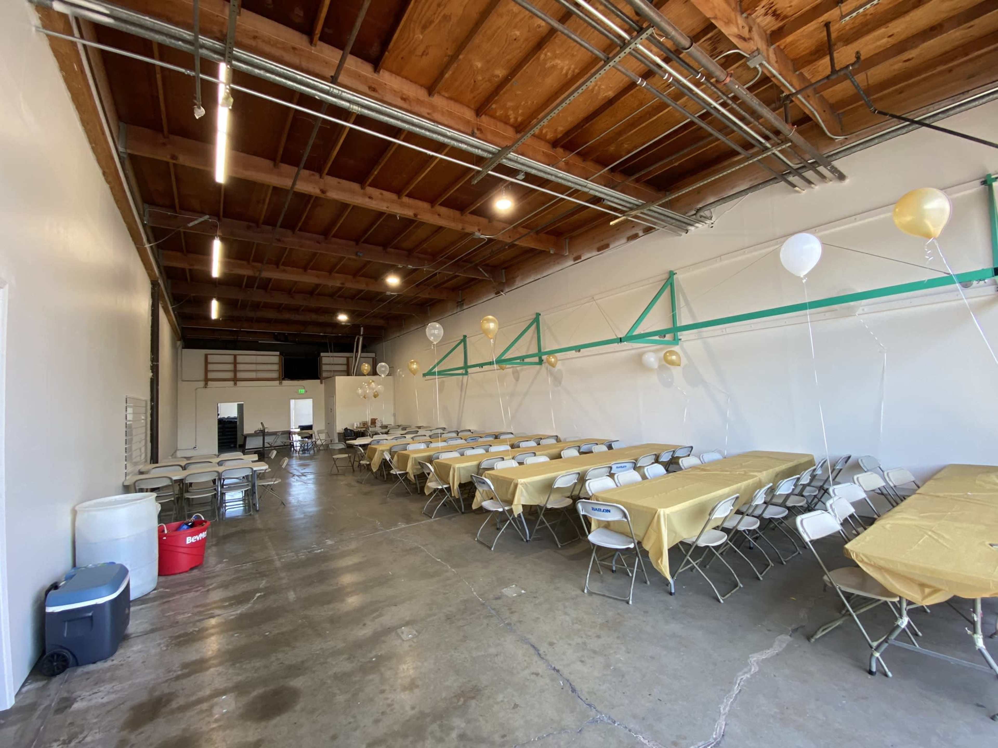 A large, empty indoor space set up with folding tables covered in gold tablecloths and surrounded by white chairs, with balloons suspended along the walls.