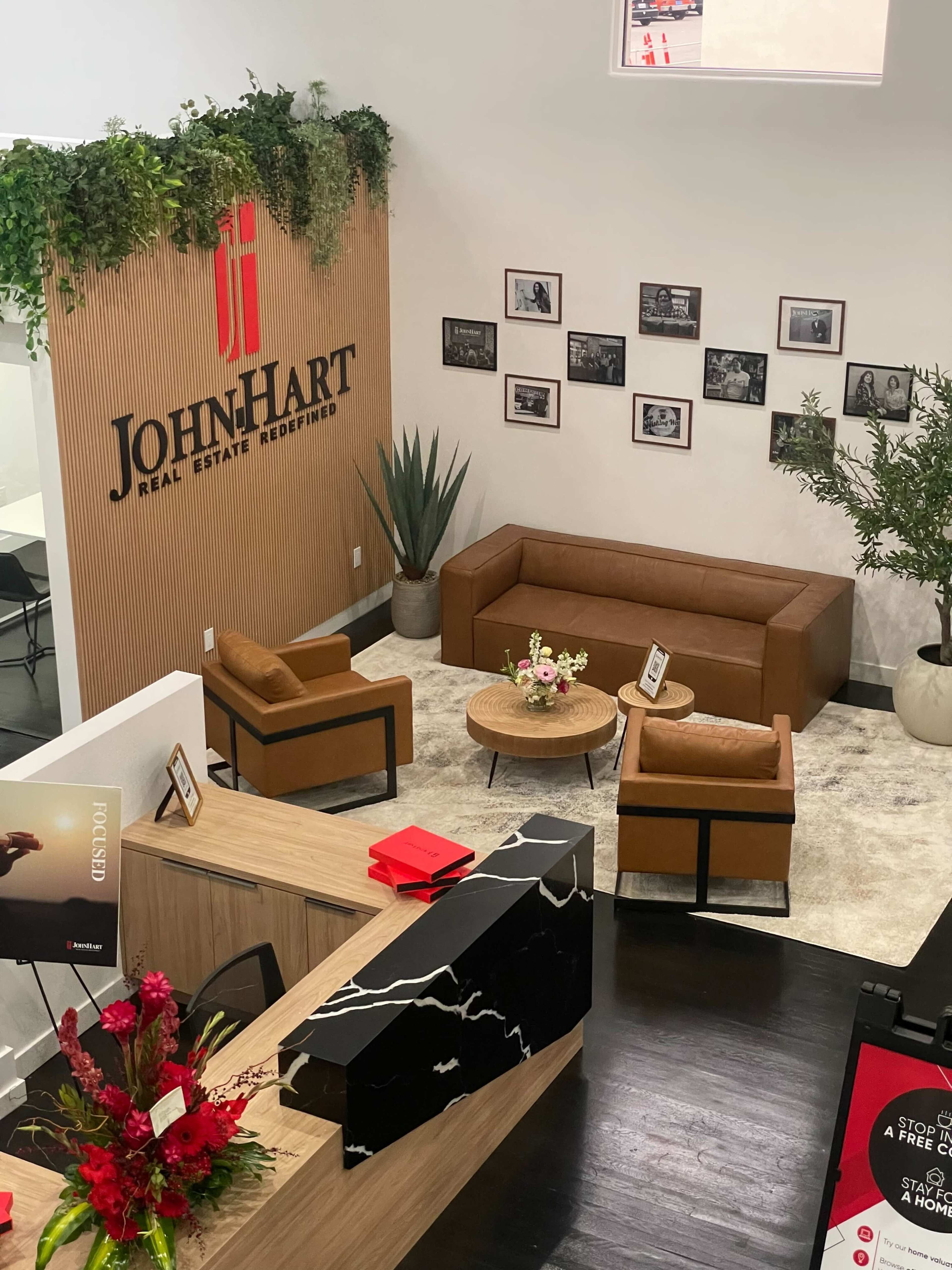 The image shows a modern office lobby with a reception desk, a seating area featuring two brown chairs and a couch, and a wall adorned with photographs and plants.