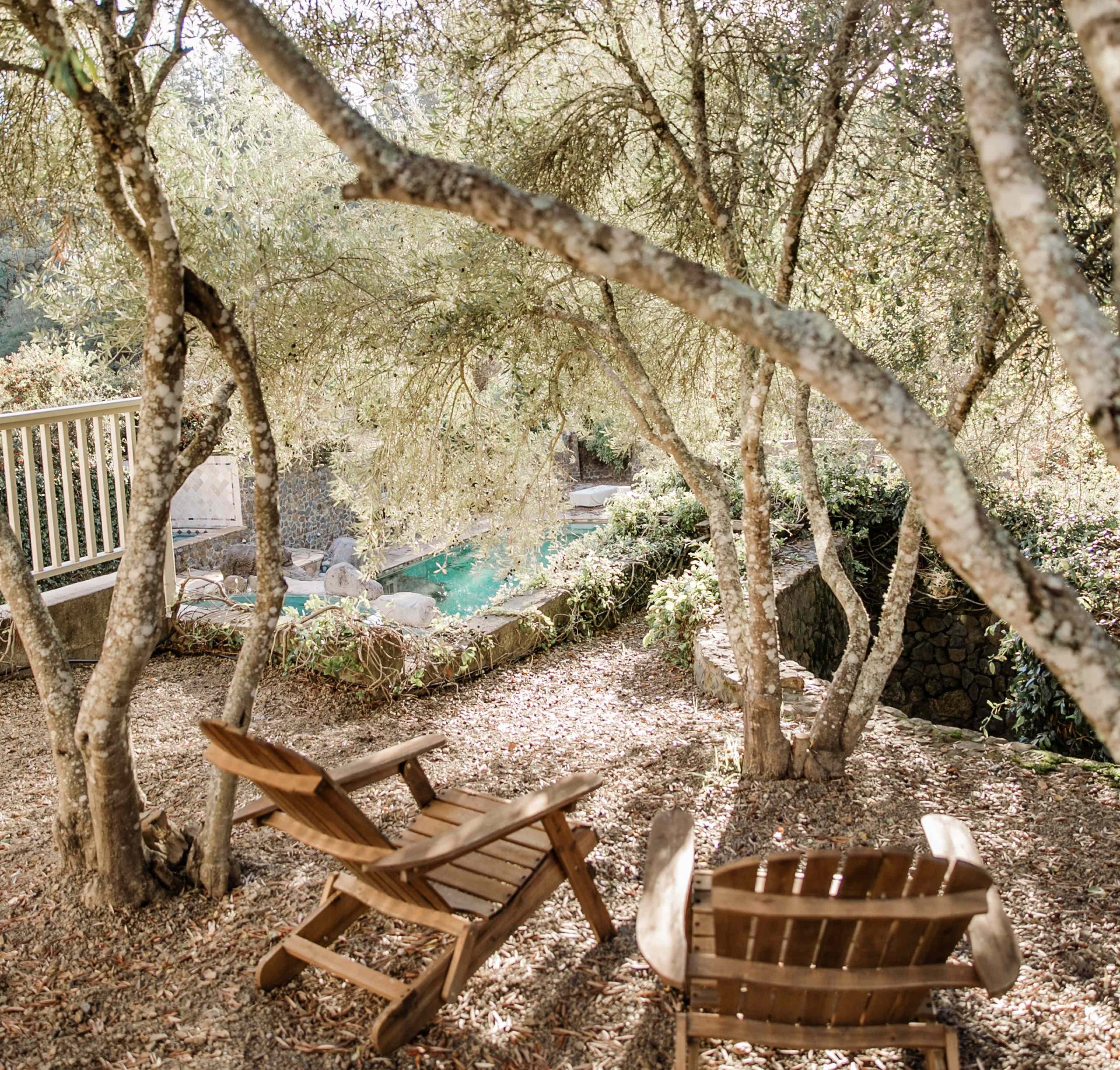 Two wooden Adirondack chairs are positioned under trees, overlooking a pool surrounded by stone walls and greenery.