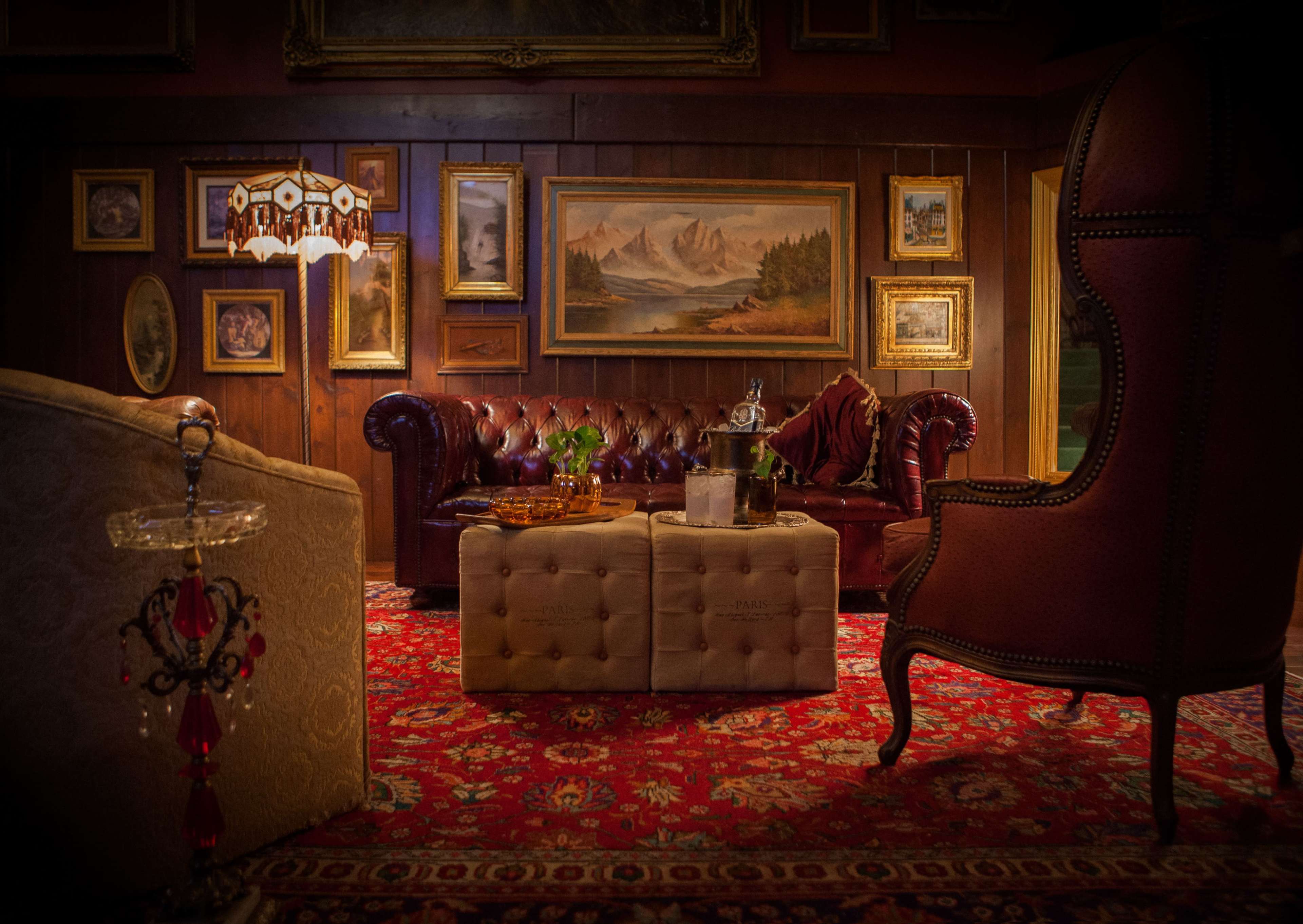 The scene features a rustic lounge with a leather couch, ornate armchair, a table with drinks, and framed artwork on wooden walls.