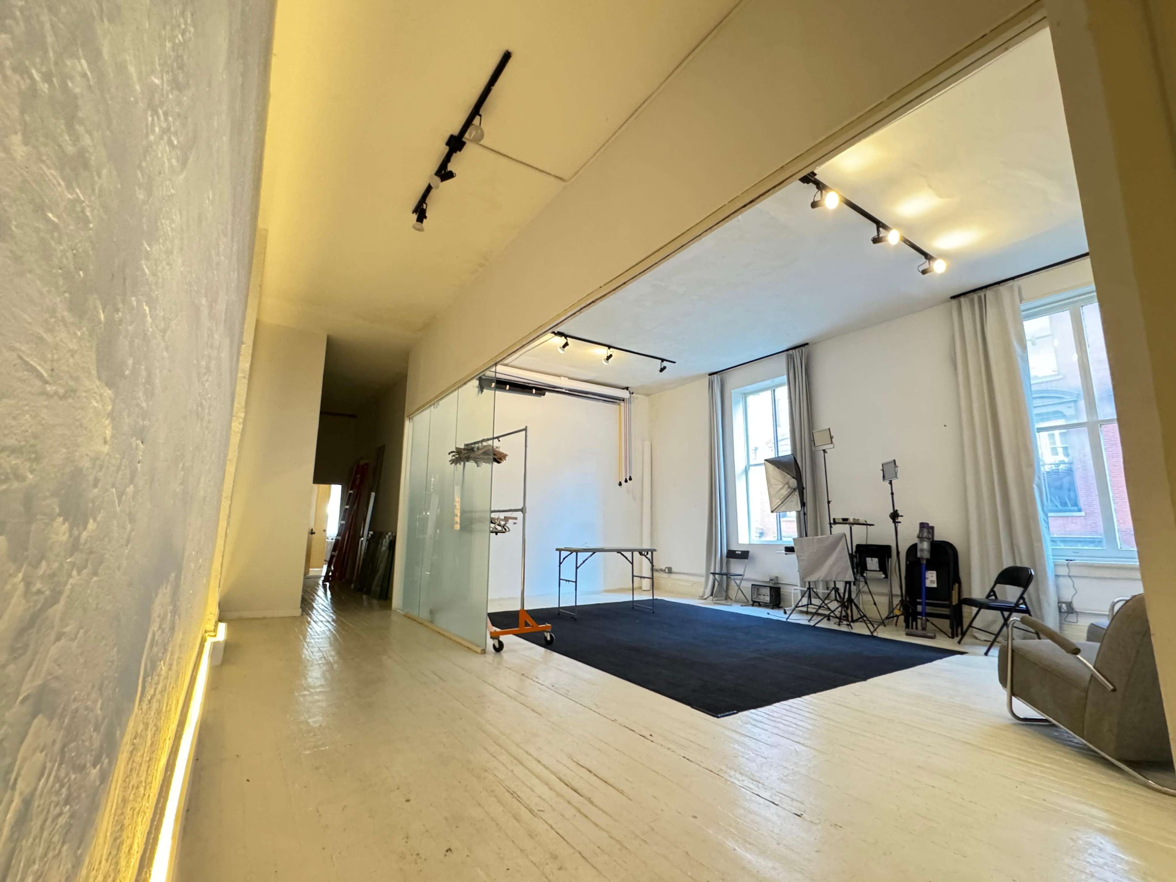 The image shows a spacious room with light-colored walls, a polished wooden floor, and a glass partition separating areas, featuring studio lighting and equipment in one section.