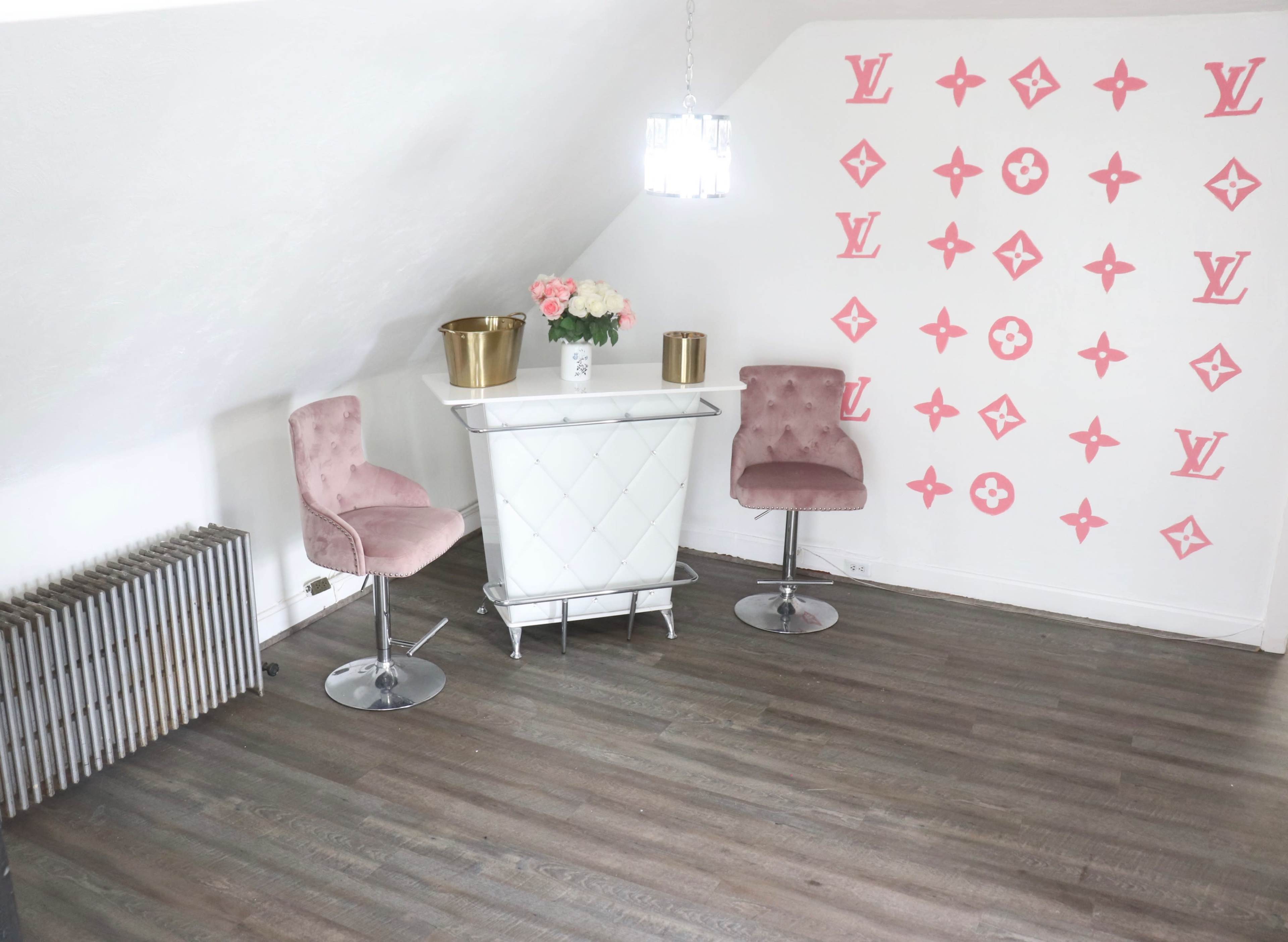 A small room with two pink stools, a white table, and a wall decorated with a repeated pattern featuring a designer logo.