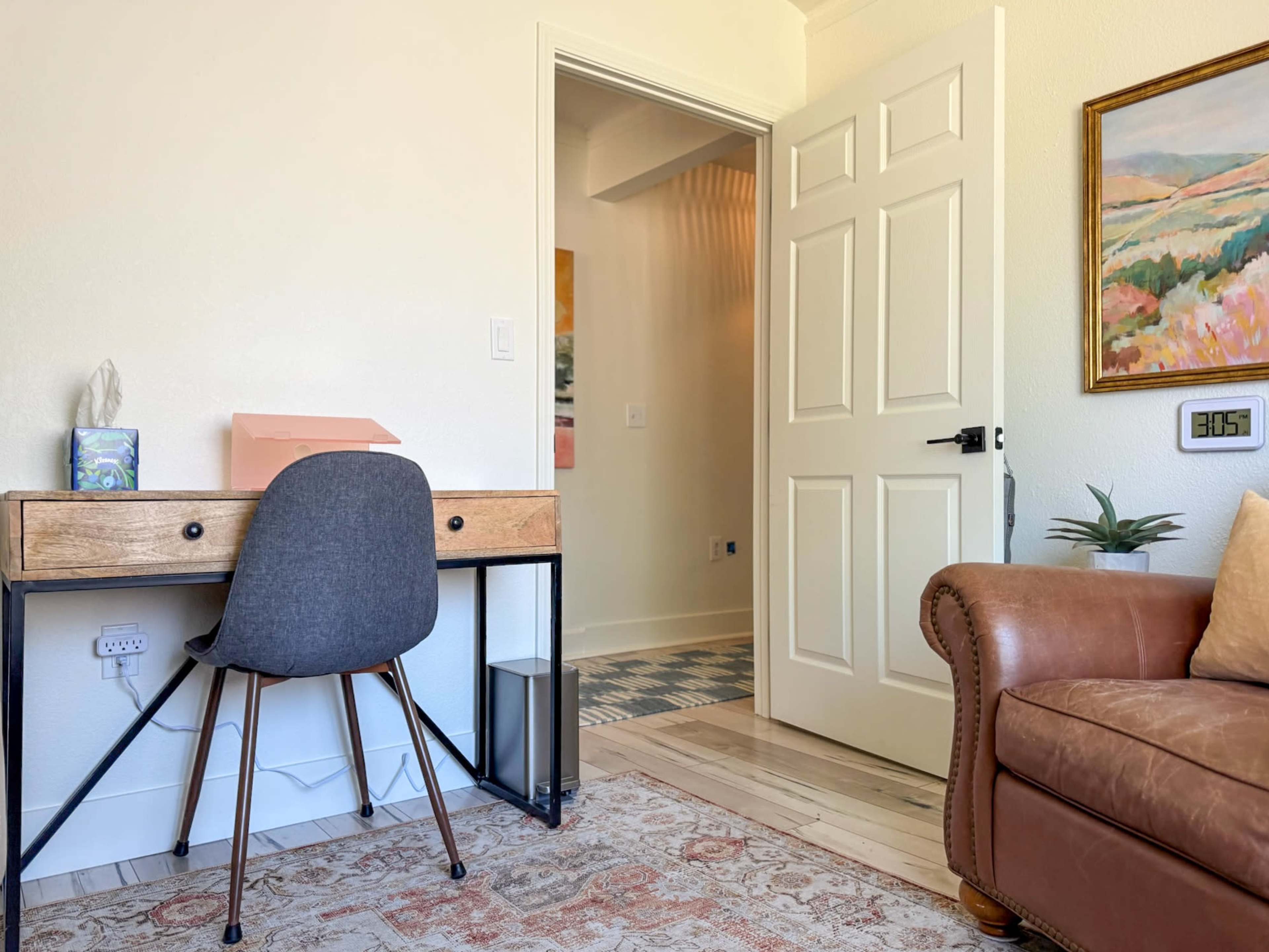 A small, well-lit room features a wooden desk with a gray chair on one side and a brown leather couch on the other, with a door leading to another area in the background.