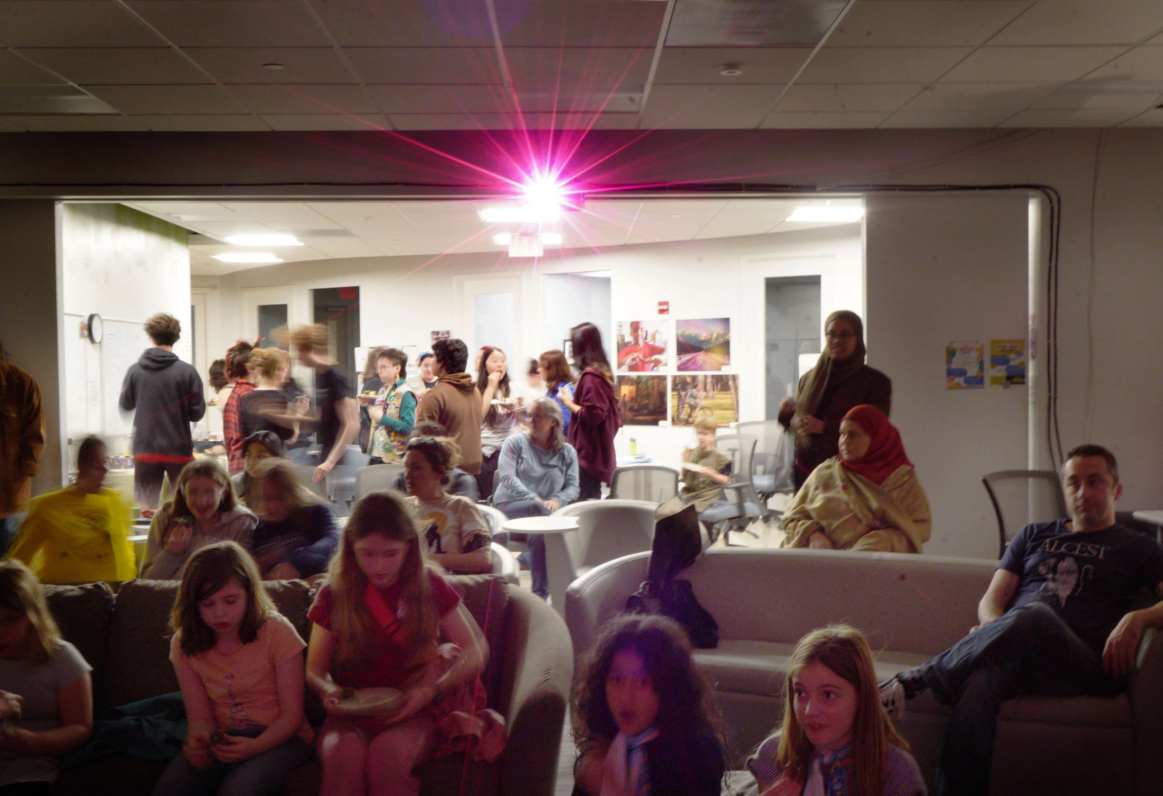 A group of people gather in a room with a large light source, while some sit on a couch and others stand or converse in the background.