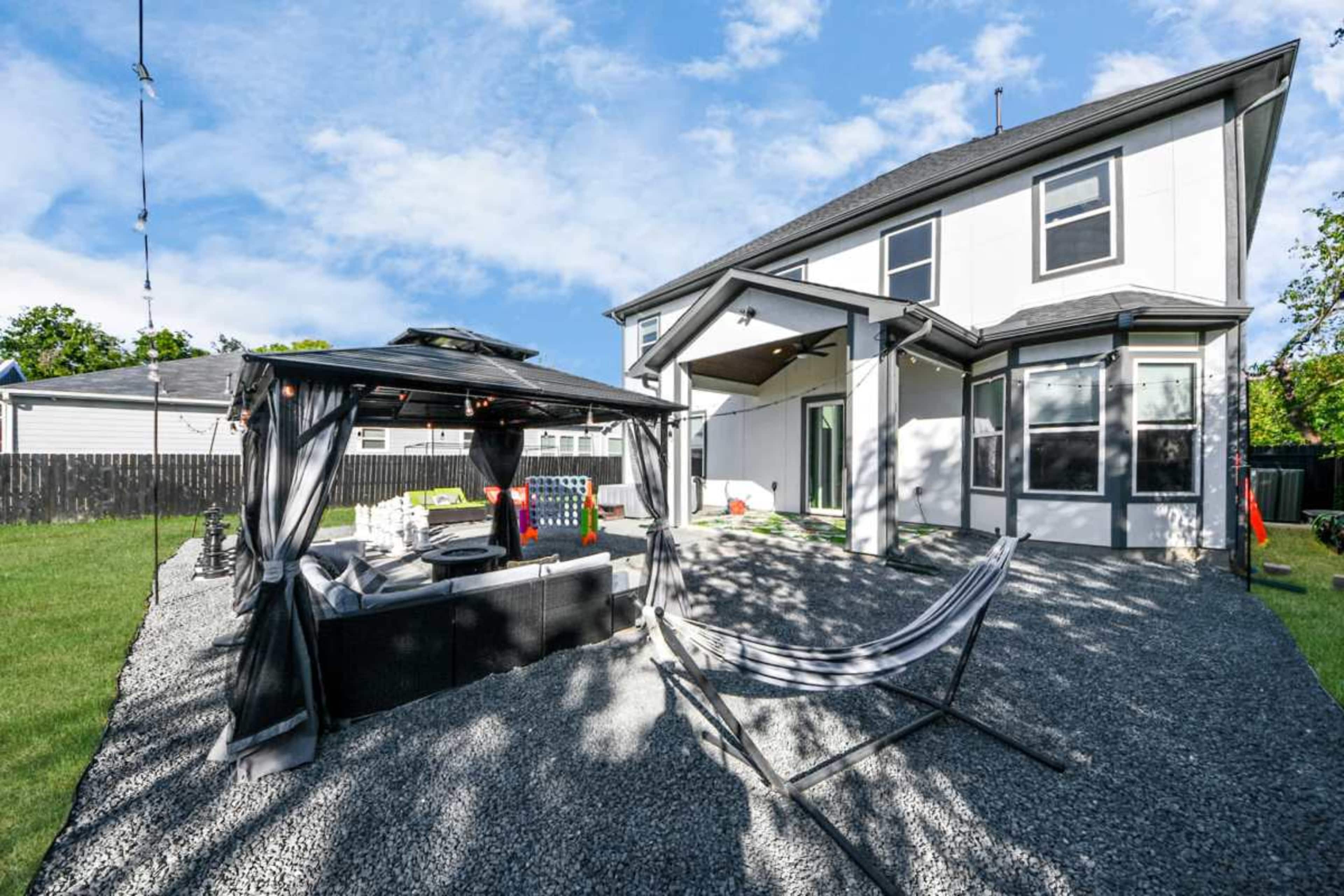 The image shows a backyard with a gravel surface, a black gazebo with seating, a hammock, and a two-story house in the background.
