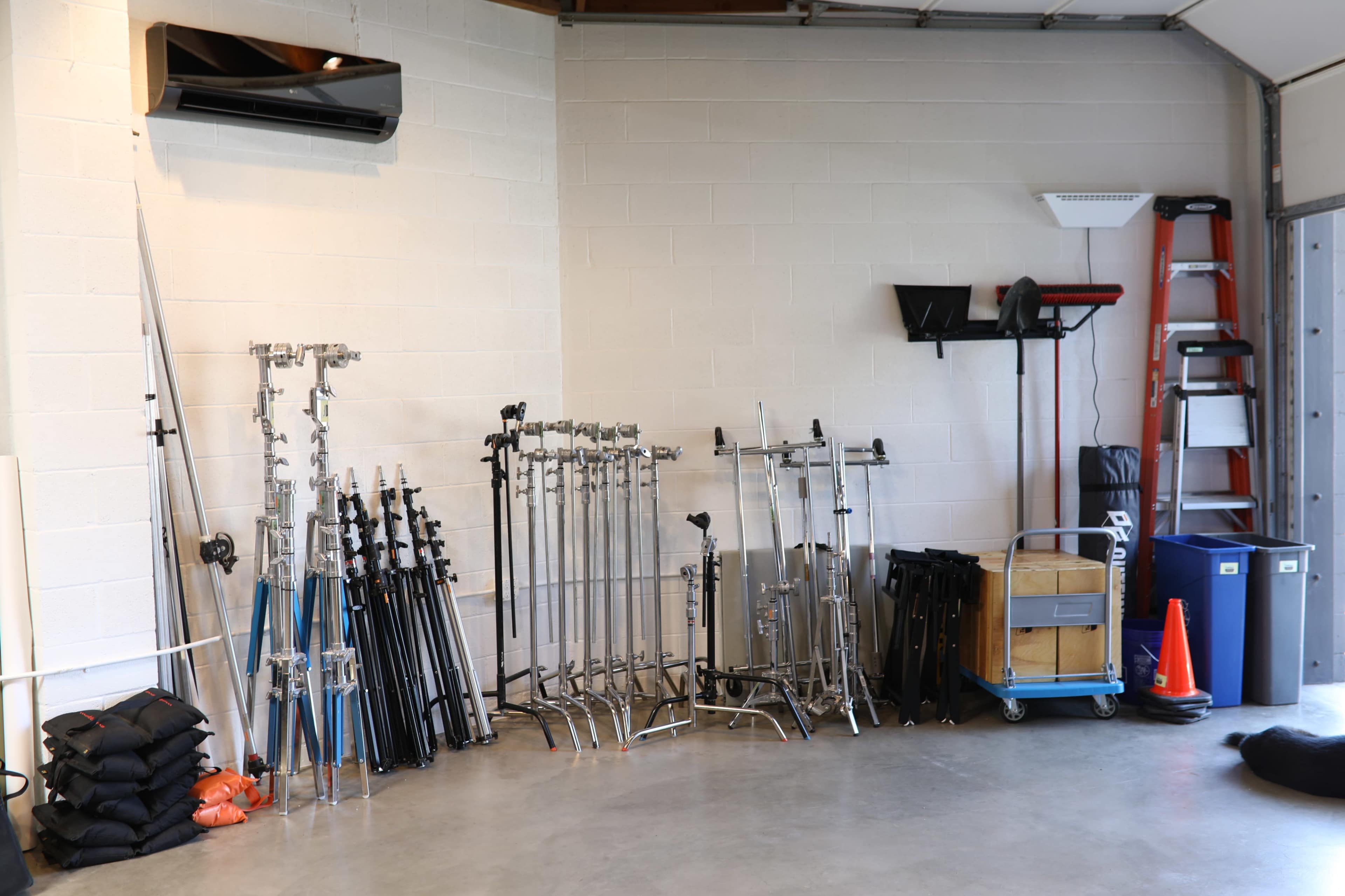 A garage space with various photography equipment, including multiple light stands, sandbags, and a cart, neatly arranged against the wall.