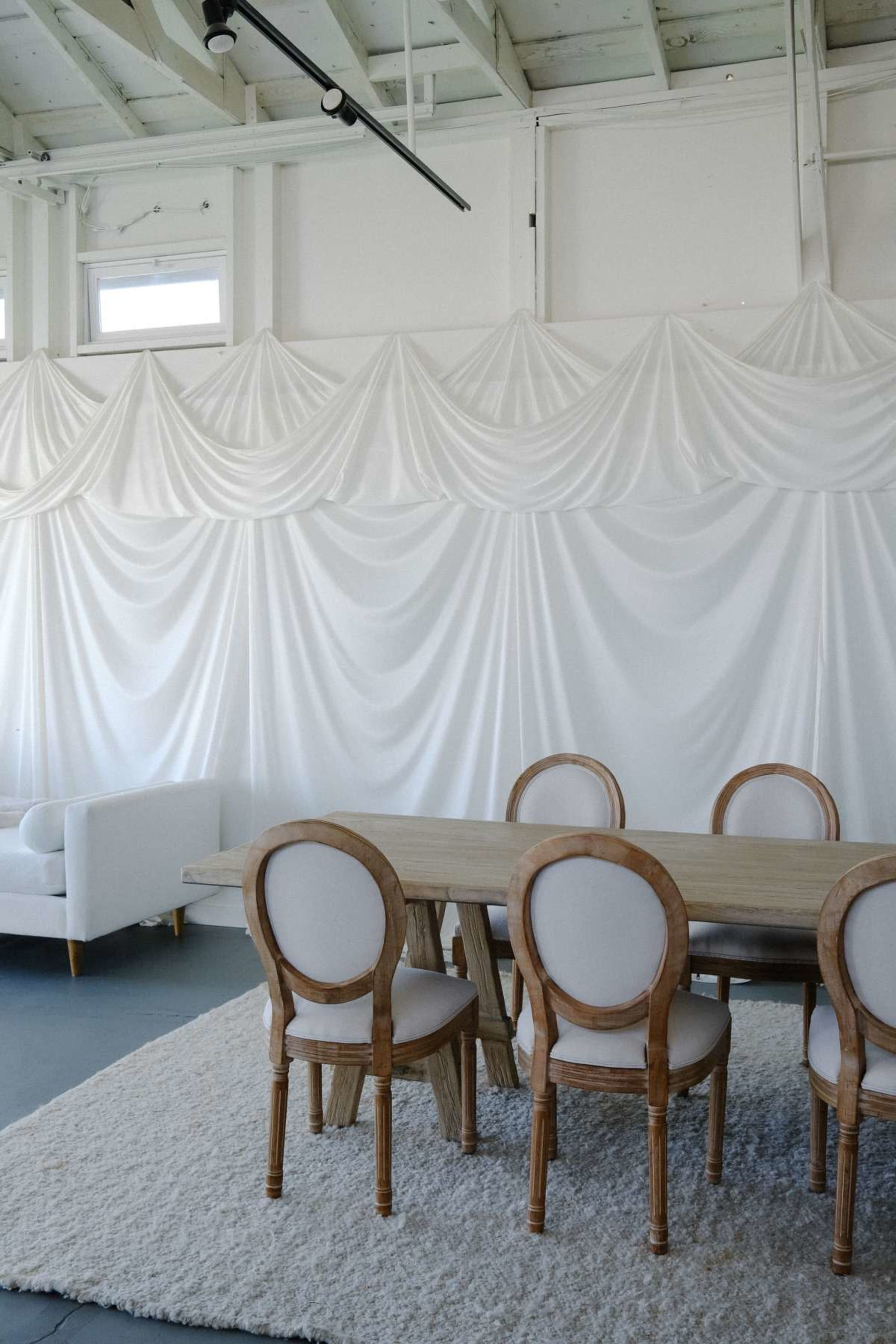 The image shows a dining area with a wooden table and six upholstered chairs set against a white draped wall.