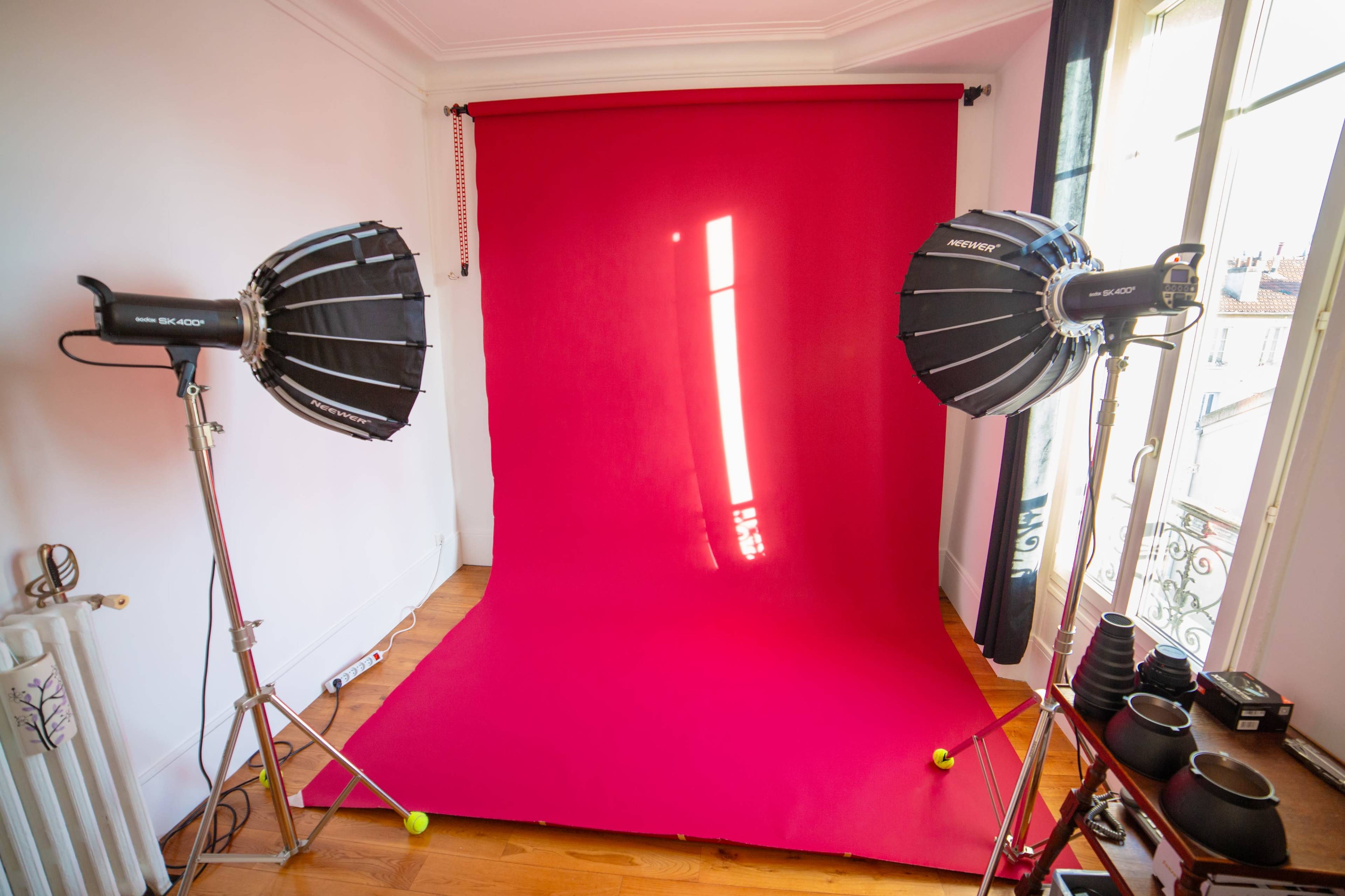 The image shows a photography studio setup with a bright pink backdrop, two light sources, and a collection of camera lenses on a shelf.